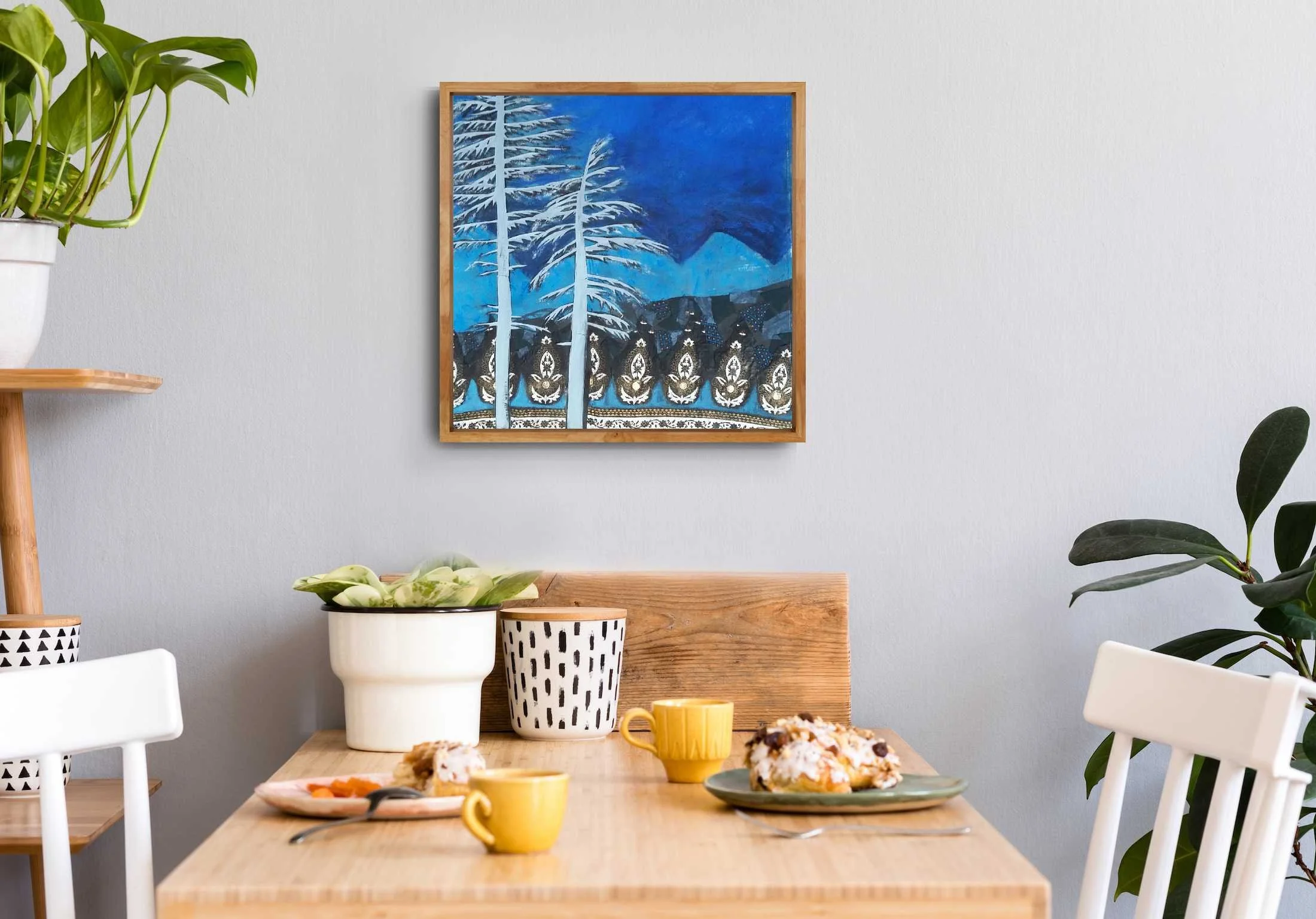 A cozy dining corner with a wooden table set for breakfast, two plates with pastries, two yellow mugs, and decorative plants on the side, with a blue landscape painting on the wall.