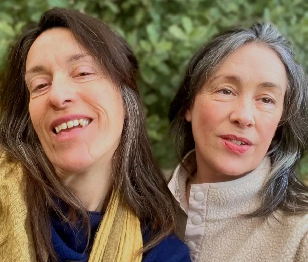 Two women with long, dark hair, one with gray streaks, posing outdoors with a green leafy background.