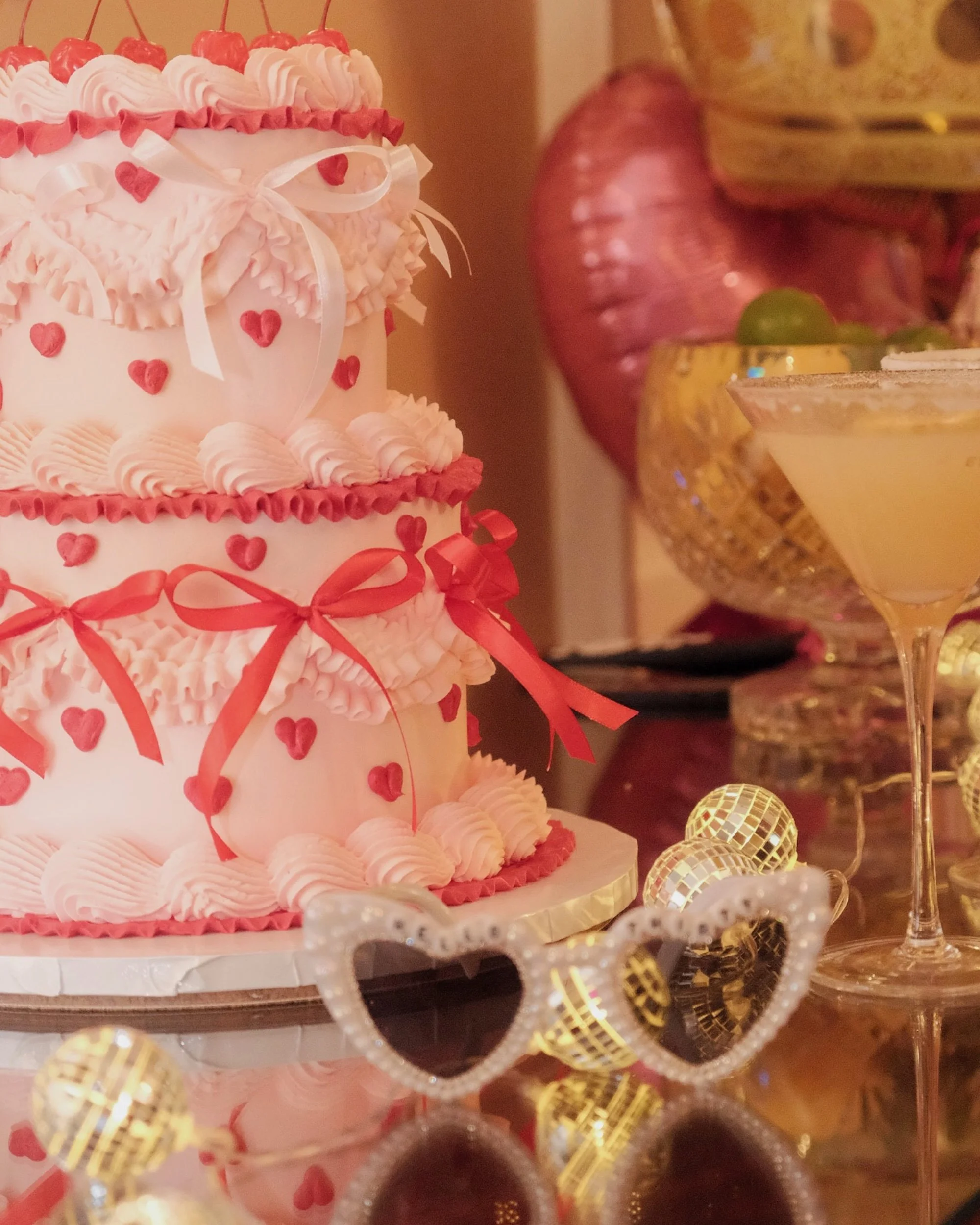 A three-tier pink and red heart-themed birthday cake with ribbons, heart-shaped decorations, and pink frosting, surrounded by gold and heart-shaped decorations, glasses, and heart-shaped sunglasses on a reflective surface.