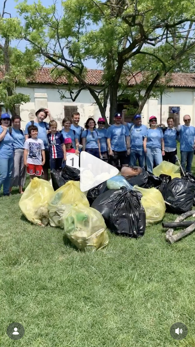 Group of people standing outdoors with bags of collected trash after a cleanup event, under a large leafy tree in front of a white building.