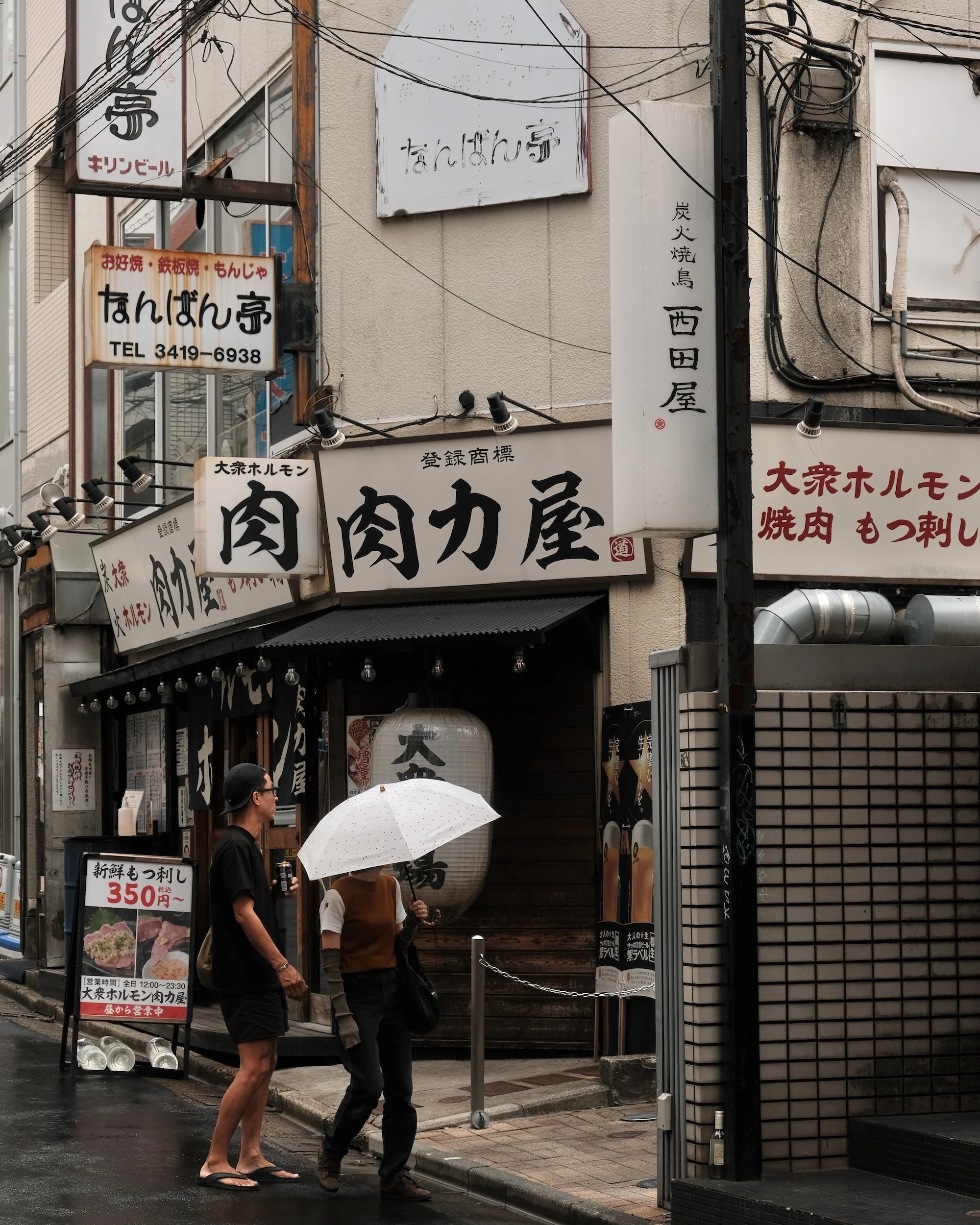 Two people walking with an umbrella past a Japanese restaurant on a street, with multiple signs and a paper lantern outside.