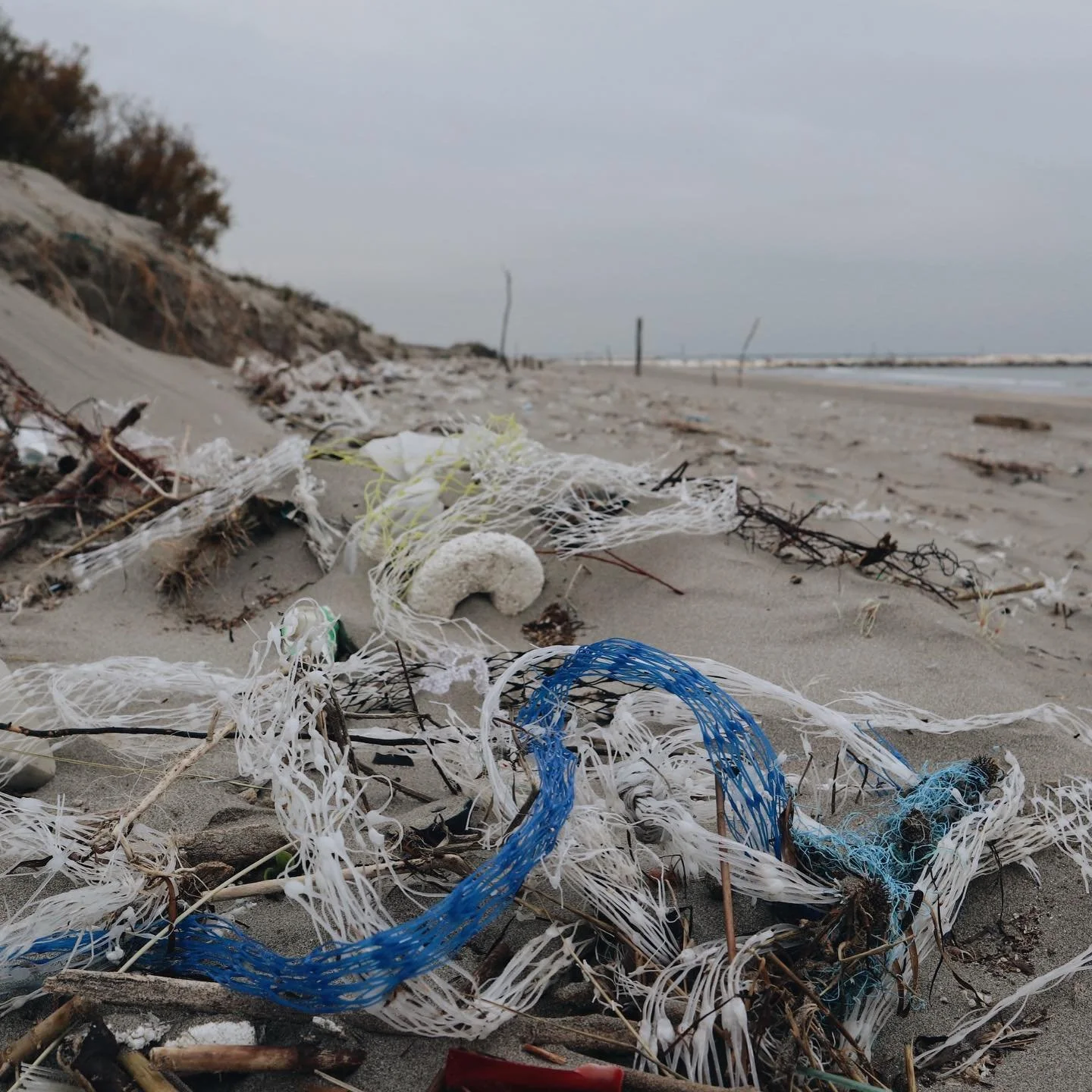 Beach with scattered trash and plastic debris, including ropes and fragments, on the sand near the shoreline with a cloudy sky.