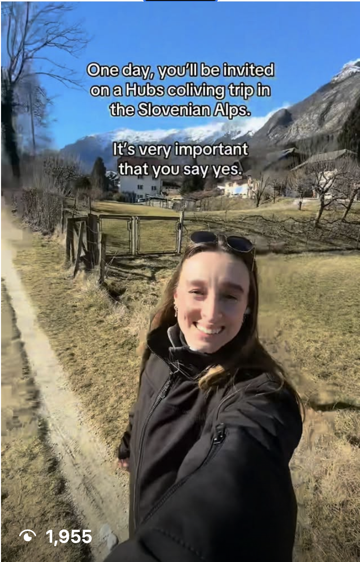 A woman taking a selfie outdoors in a rural area with mountains in the background. The sky is clear and blue. Text overlay reads, "One day, you'll be invited on a Hubs colving trip in the Slovenian Alps. It's very important that you say yes."