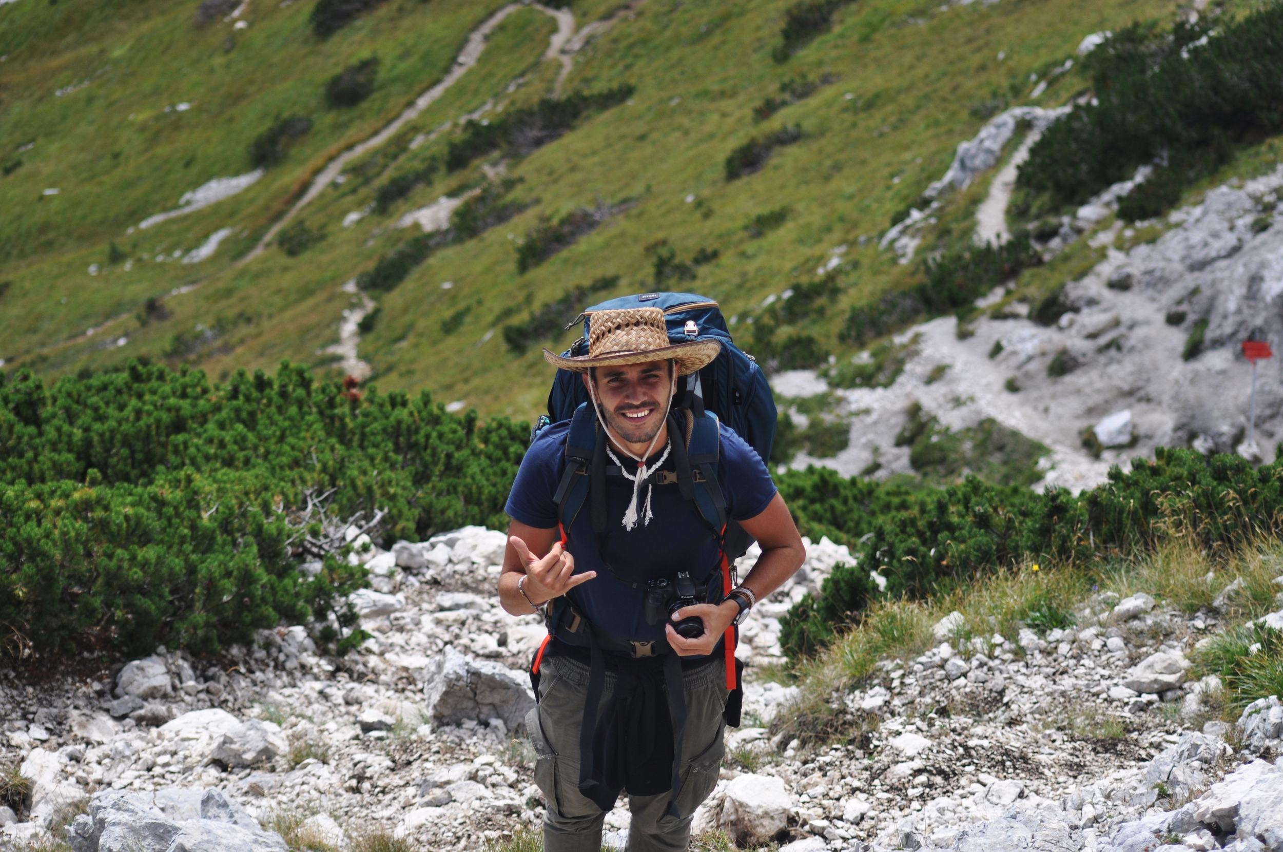 Un homme souriant en randonnée en montagne, portant un chapeau en paille, un grand sac à dos, et tenant un appareil photo, sur un sentier rocheux avec des collines verdoyantes en arrière-plan.