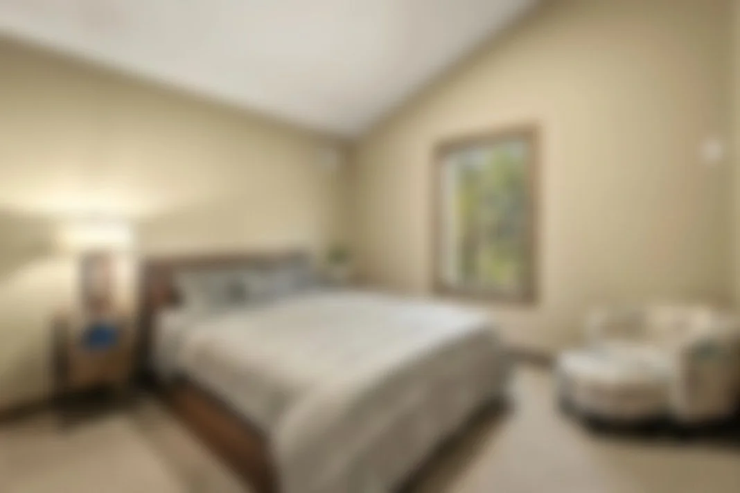 A bedroom with a bed, nightstand with lamp, decorative pillow, window showing trees outside, and an armchair with a swirl pattern.