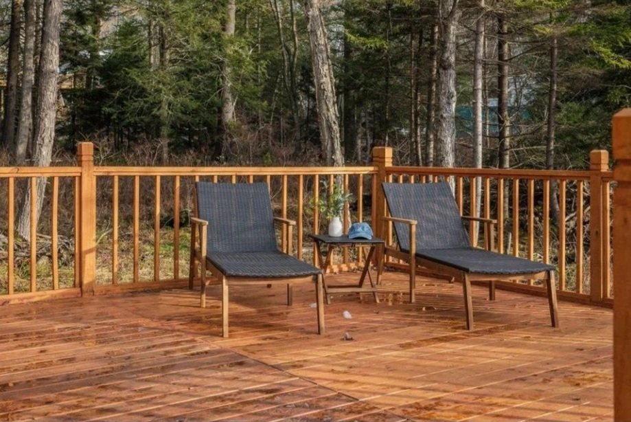 An outdoor wooden deck with two lounge chairs, a small side table, and a forest background.