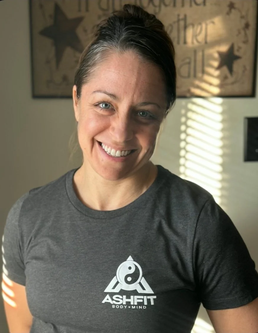 A woman with brown hair pulled back, smiling, wearing a dark gray ASHFT t-shirt with a yin-yang logo, in a room with a decorative wall hanging and sunlight coming through blinds.