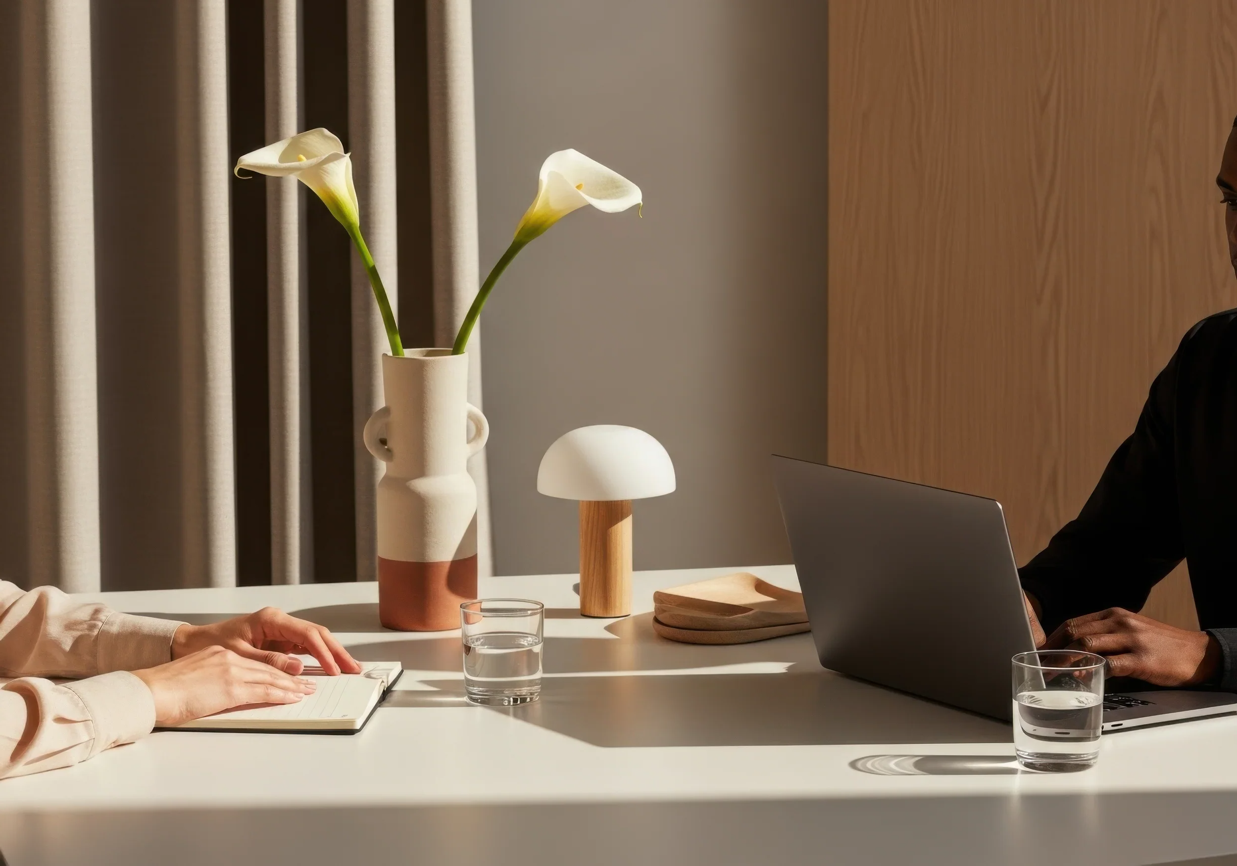 A meeting with two people at a white table, one with hands on a notebook, the other typing on a laptop, with a vase of white calla lilies, a small mushroom-shaped lamp, and two glasses of water on the table.
