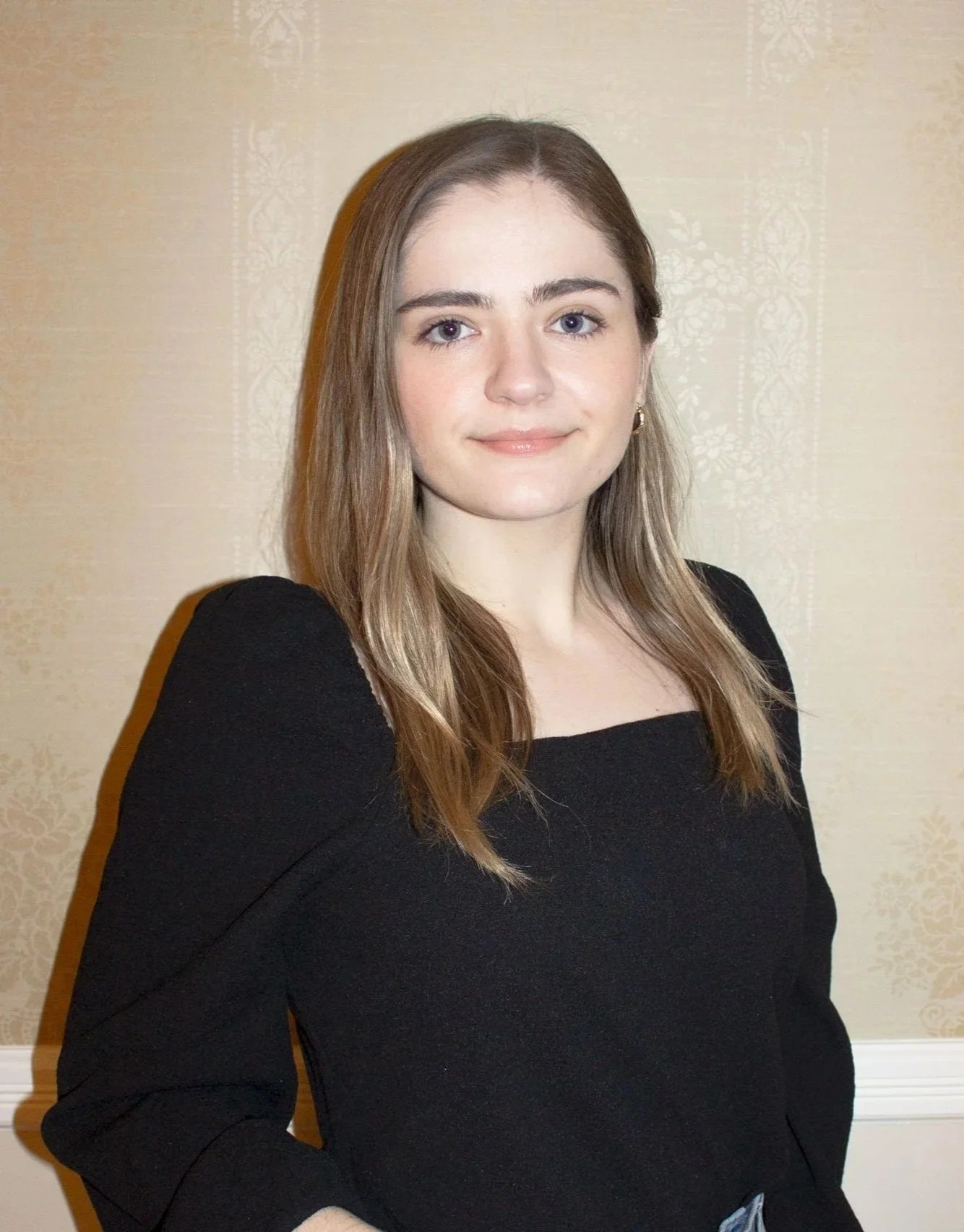 A young woman with long, light brown hair and fair skin wearing a black top standing against a patterned beige wall.