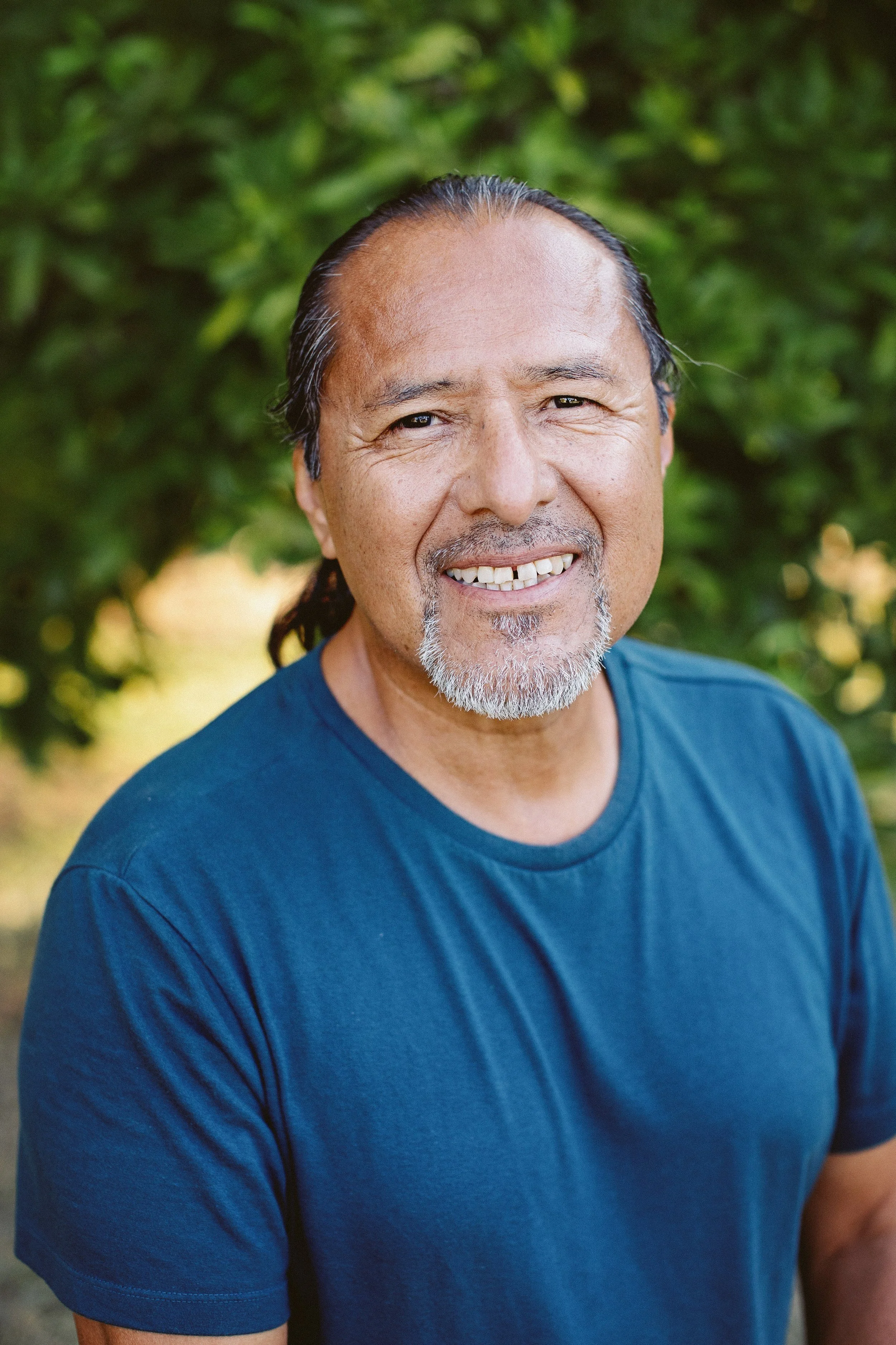 Close-up of a smiling middle-aged man with long gray hair, light mustache and goatee, wearing a blue T-shirt, with green foliage background.