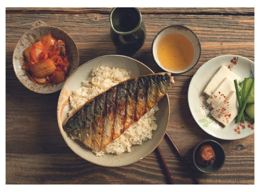 A wooden table set with a bowl of rice topped with grilled fish, a bowl of kimchi, a cup of tea, and a plate of sliced tofu with green onions and cucumbers. Chopsticks and a small dish with a pickled plum are also visible.