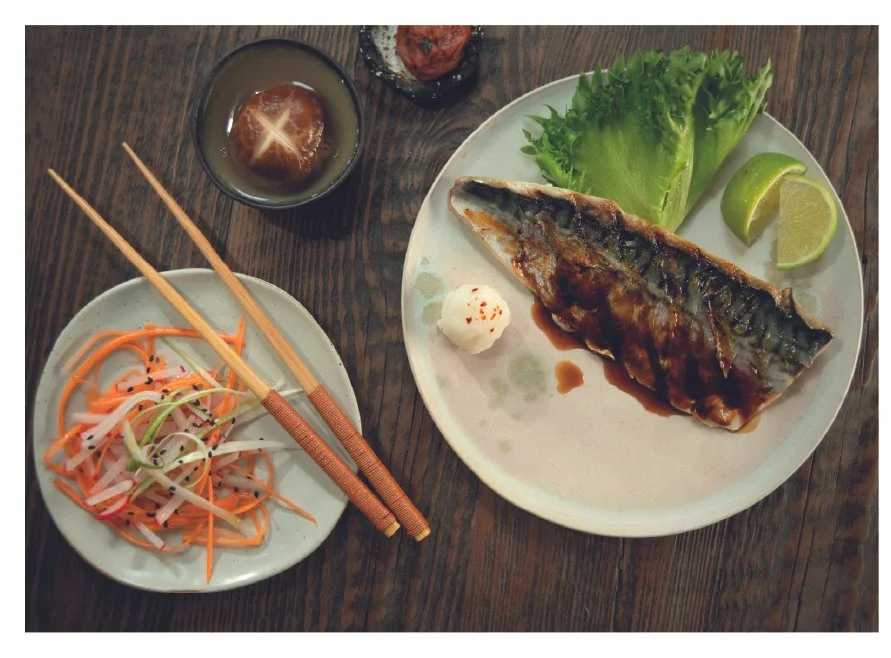 Grilled mackerel with soy sauce glaze on a plate with lettuce and lime; side of pickled vegetables and chopsticks; shiitake mushroom in a bowl; umeboshi on a small dish.