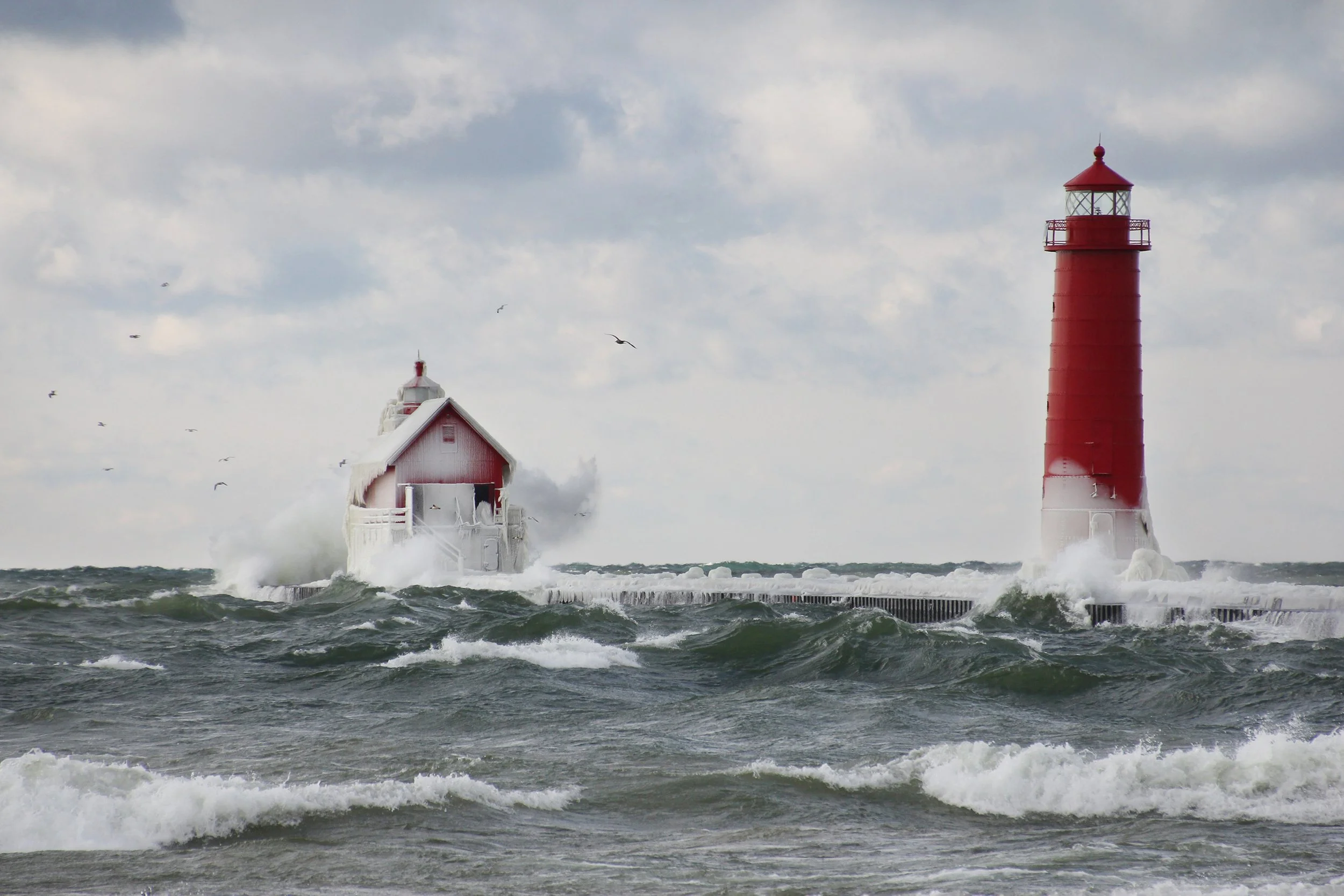 Red lighthouse and ice-covered building on a pier during rough seas with waves crashing and cloudy sky.