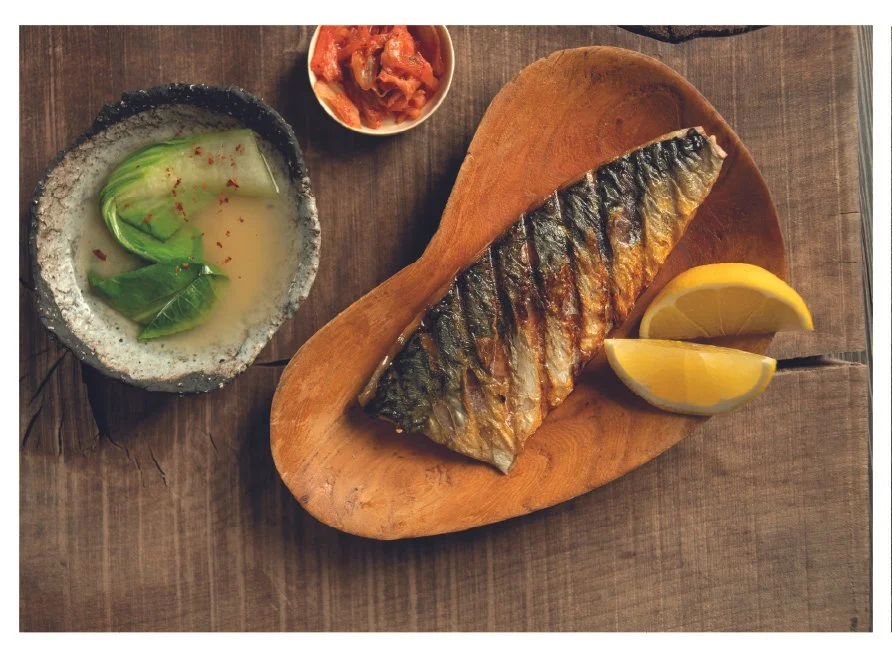 Grilled mackerel on a wooden platter with lemon wedges, small dishes of kimchi, and a green vegetable soup on a wooden table.