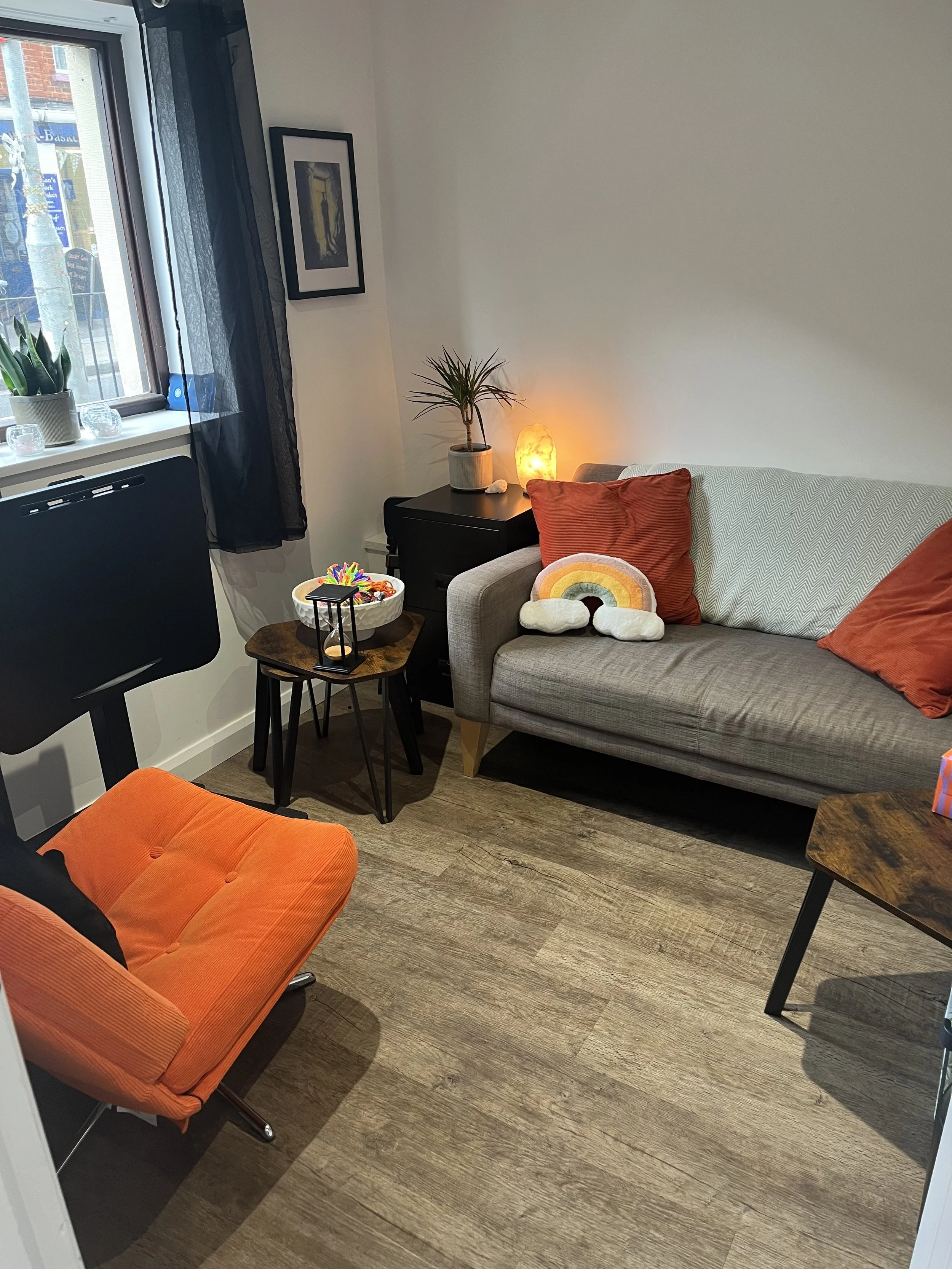A cozy therapy room with a grey sofa, orange throw pillows, a rainbow plush pillow, a small black side table with a potted plant, salt lamp, and decorative items, a black chair with an orange cushion, a window with black curtains, and a wooden floor.