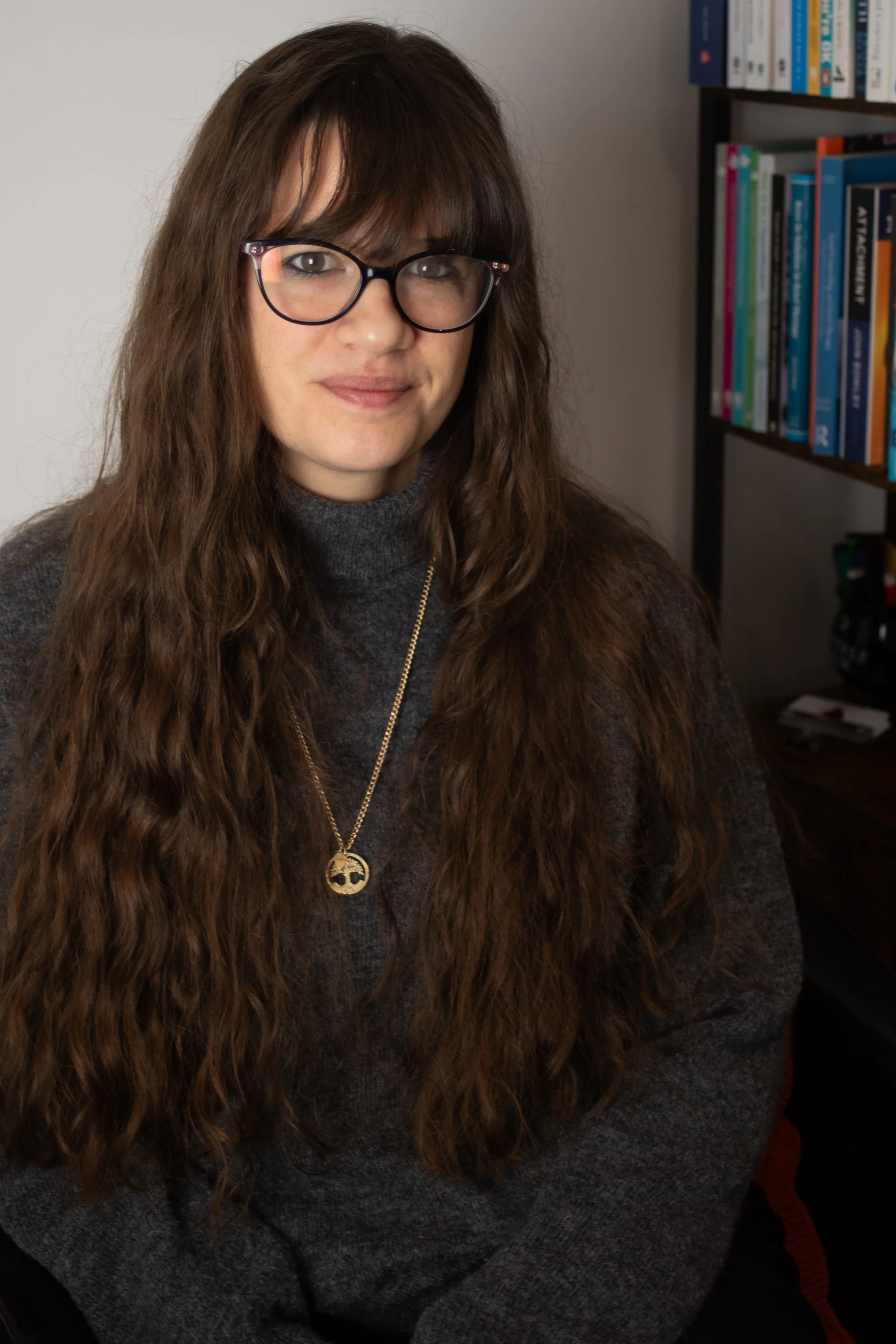 A photo of Zanny Jode, Psychotherapist with long brown hair, wearing a Grey Jumper and a gold tree of life necklace smiling at the camera against in her therapy room at Enchanted Circle, Attleborough, Norfolk. .