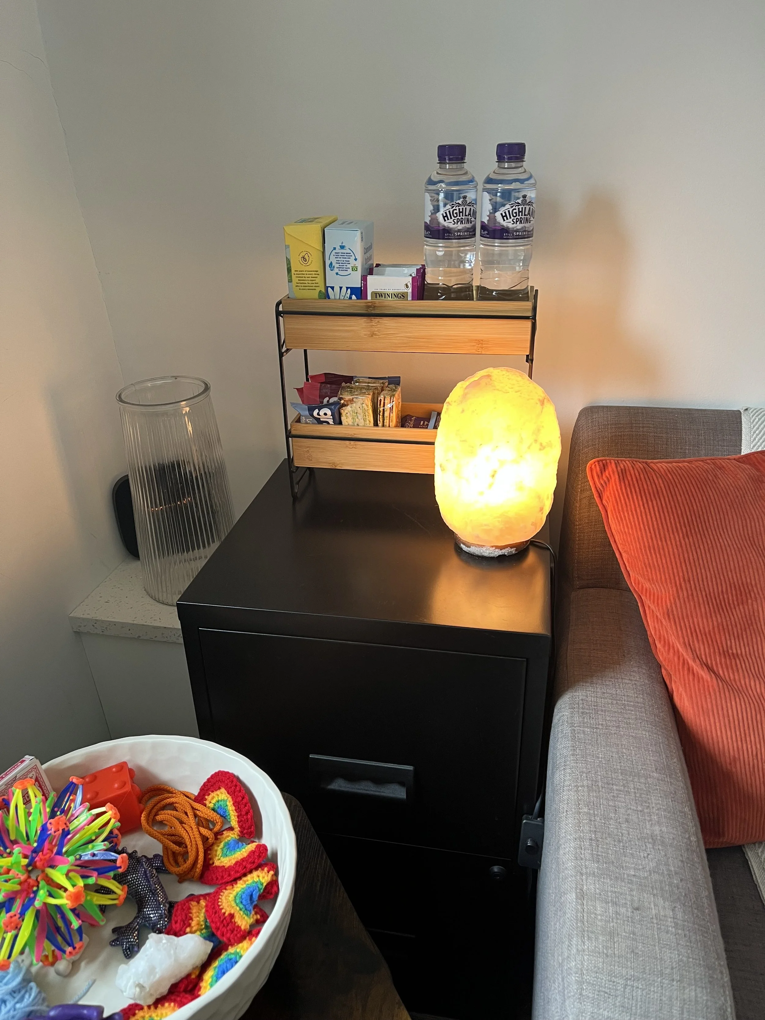 Side table with water bottles, snack boxes, and a salt lamp, adjacent to a gray sofa with a red pillow, and a plastic container filled with colorful yarn and craft supplies.