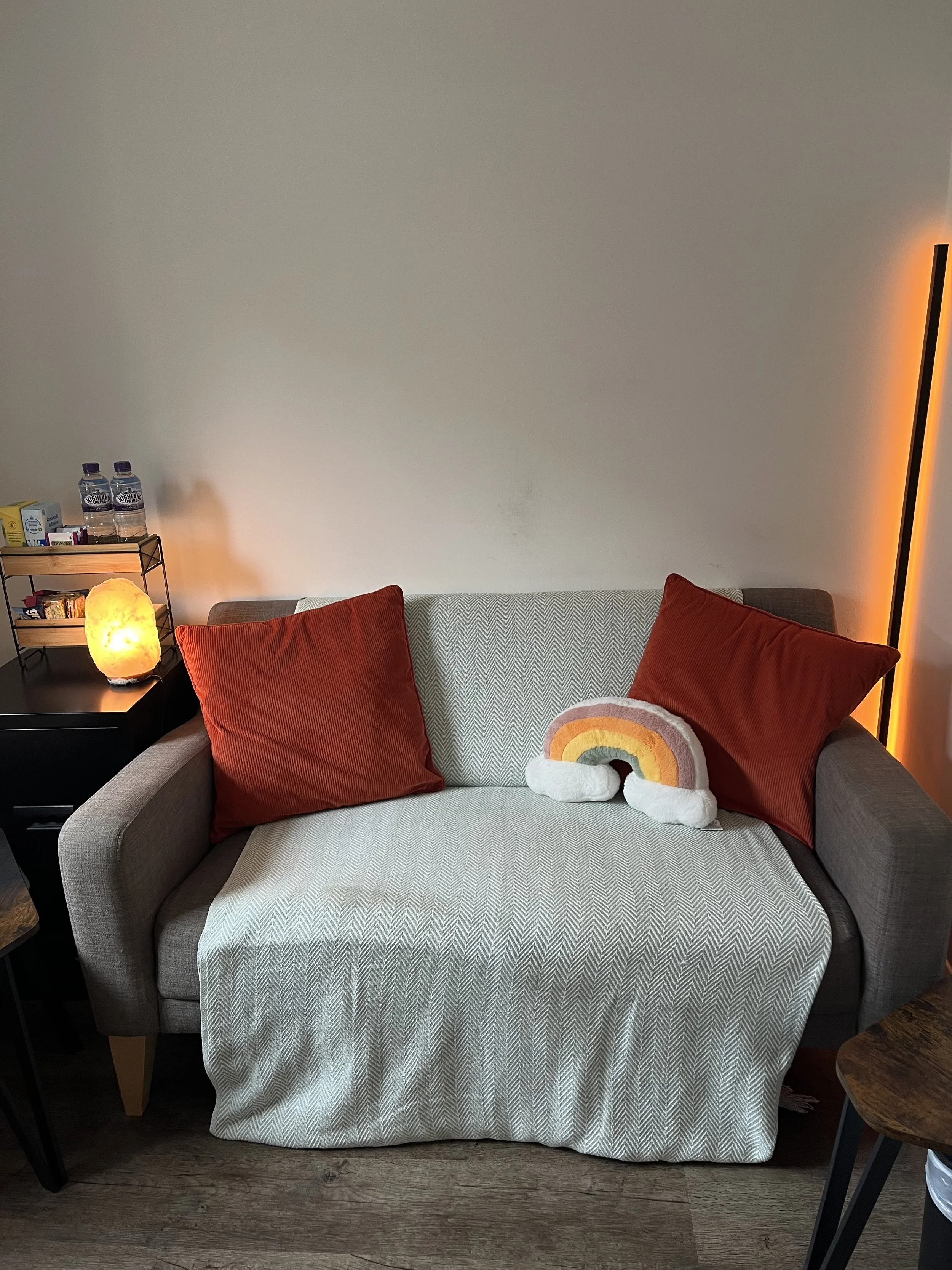 Therapy room with a gray sofa, red pillows, a white and rainbow-colored plush toy, a black side table with a salt lamp, and a corner shelf with water bottles and various items. A floor lamp with an orange light is visible on the right.