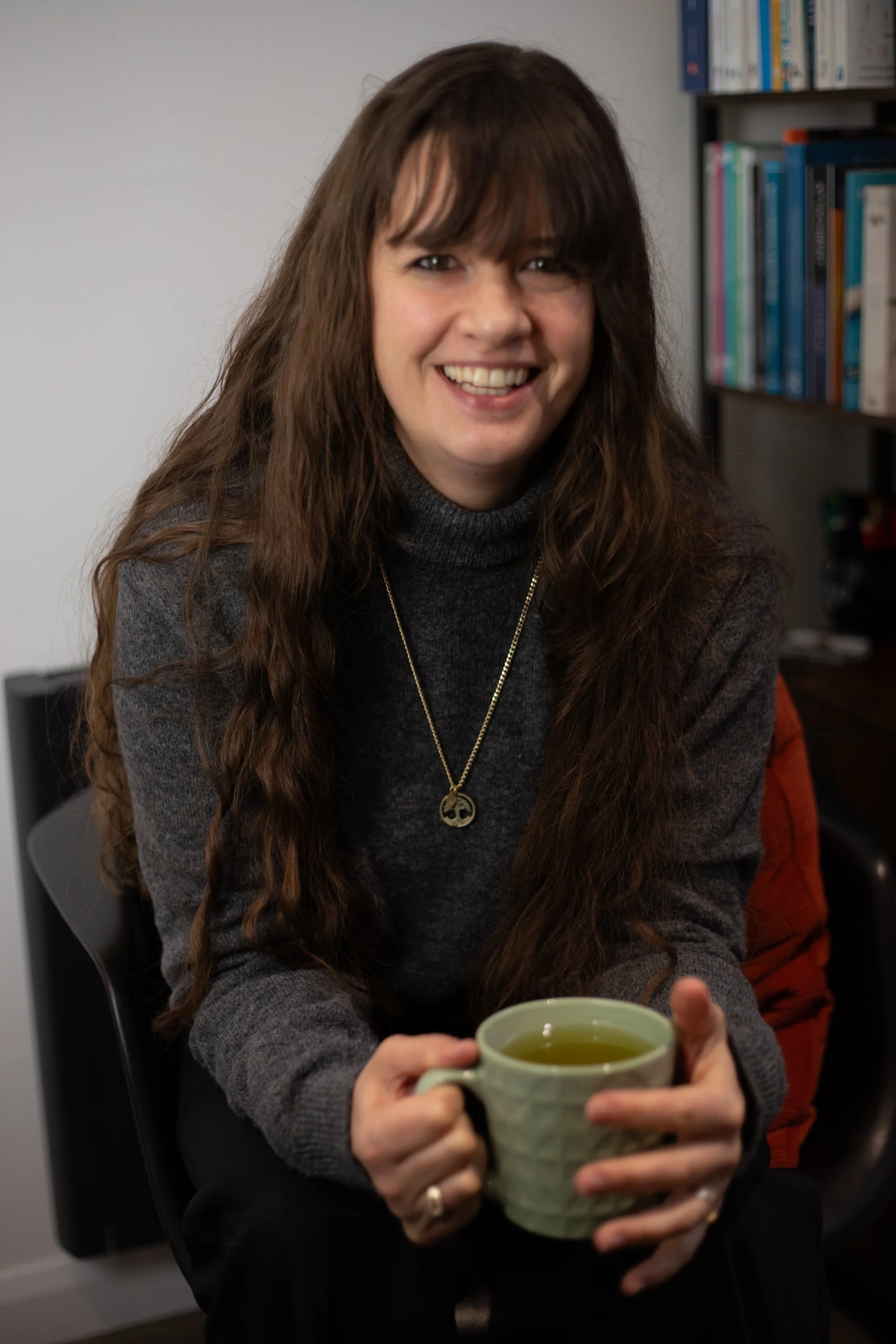 A photo of Zanny Jode, Psychotherapist drinking a hot drink, with long brown hair, wearing a grey jumper and a gold tree of life necklace smiling at the camera against a light background at Enchanted Circle, Attleborugh, Norfolk.