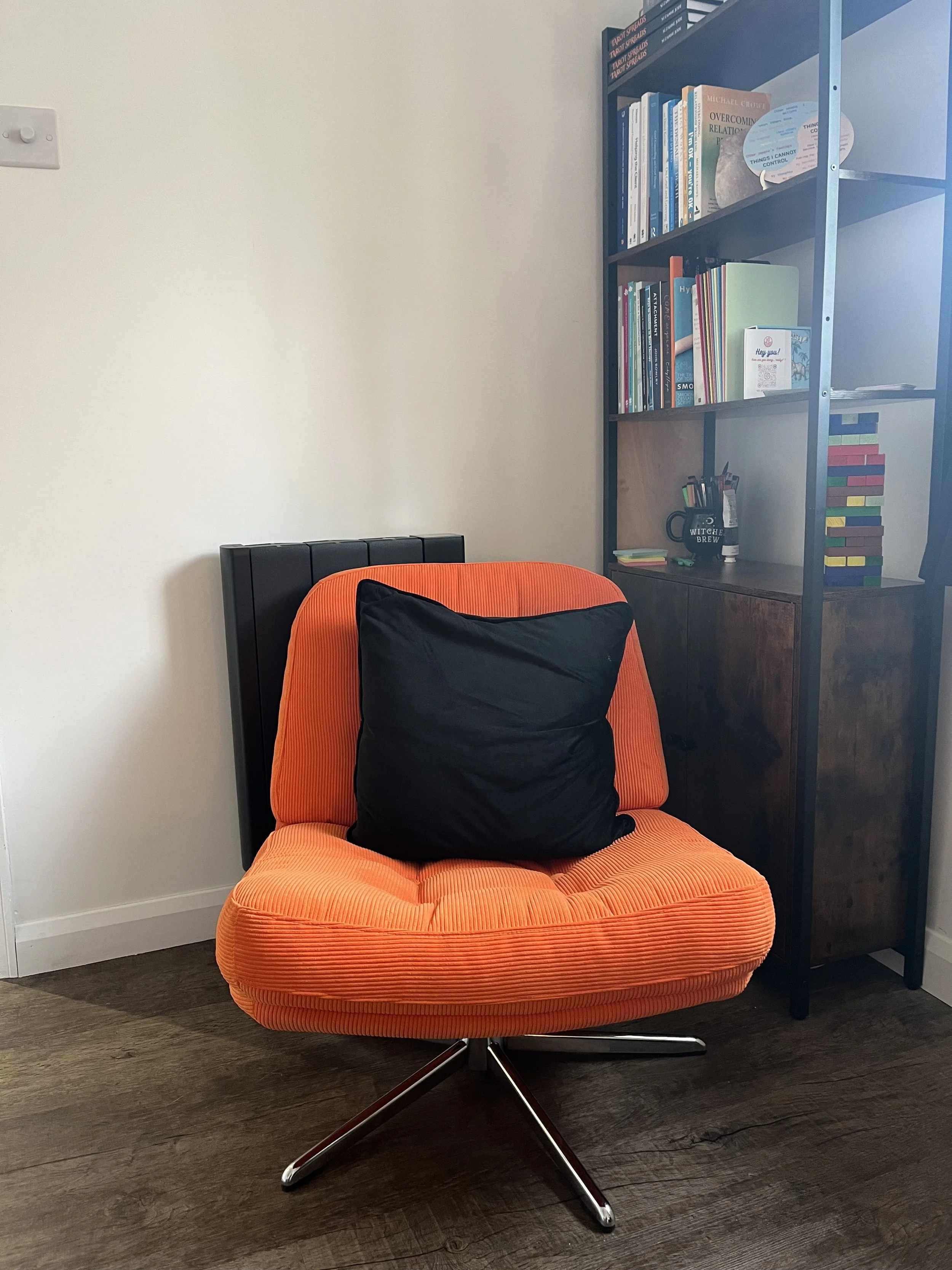 Orange swivel chair with a black cushion in front of a bookshelf with books, a mug that says 'WITCH BREW,' some pens, and a book titled 'Hey you!' in a room with wood flooring and a white wall.