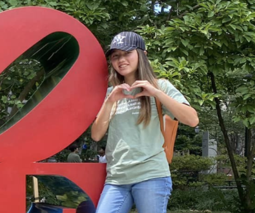 Person wearing a cap and green t-shirt making a heart shape with hands in front of a large red sculpture outdoors, surrounded by greenery.