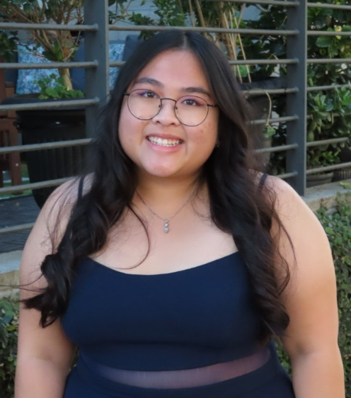 Smiling woman with long dark hair and glasses wearing a navy blue sleeveless dress, standing outdoors in front of a fence with greenery.