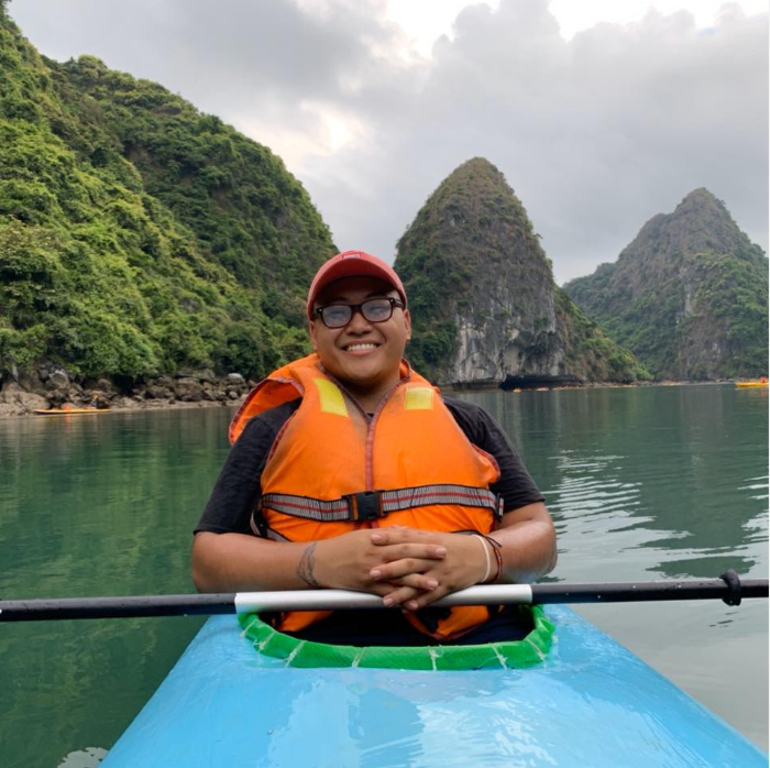 Person kayaking in calm water, wearing an orange life jacket and cap, with rocky green hills in the background.