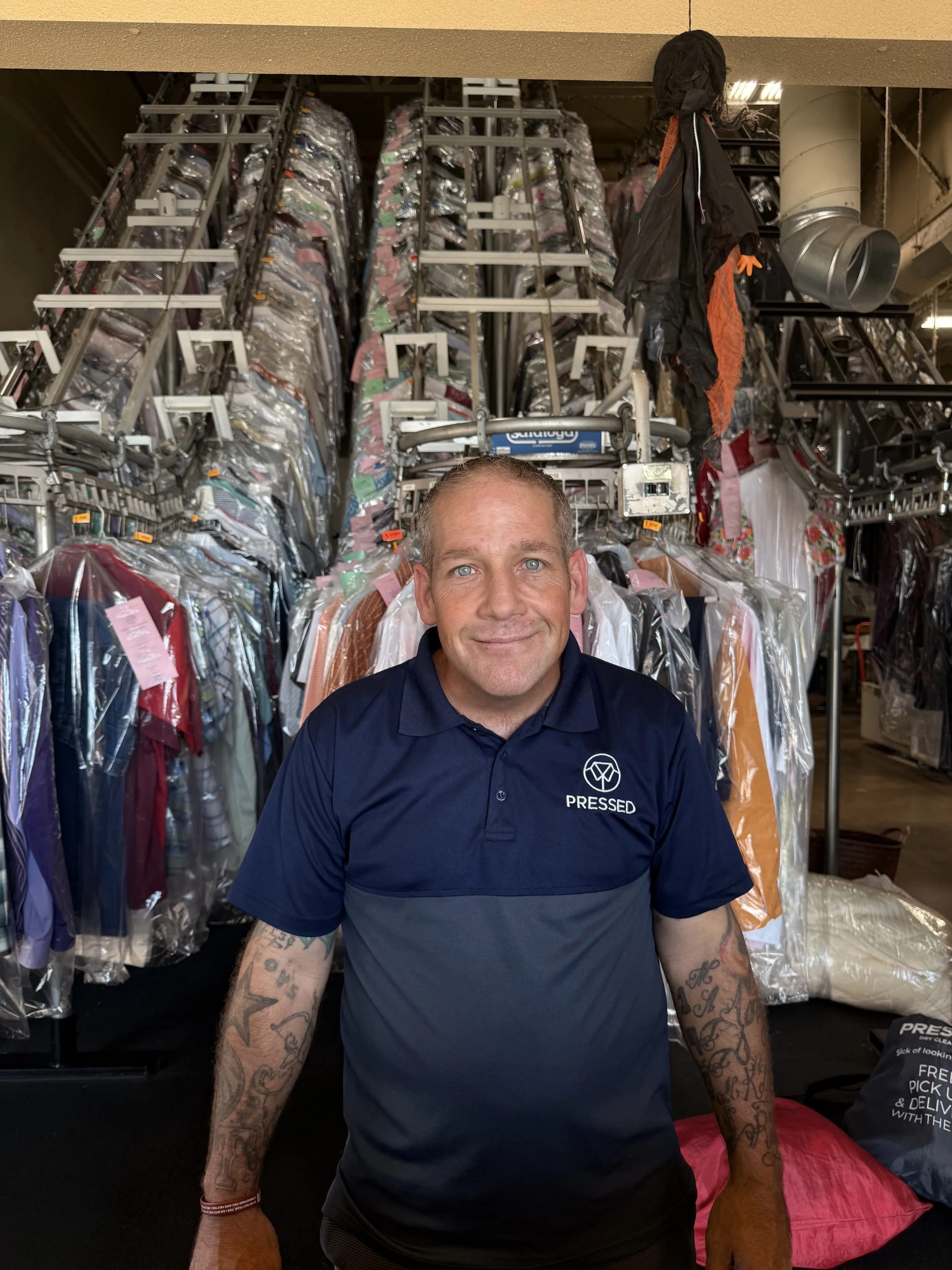 A smiling man with tattoos on his arms, wearing a navy blue polo shirt with an logo and the word 'Pressed', standing in front of clothing racks filled with hanging garments in plastic covers at a dry cleaning or laundry facility.