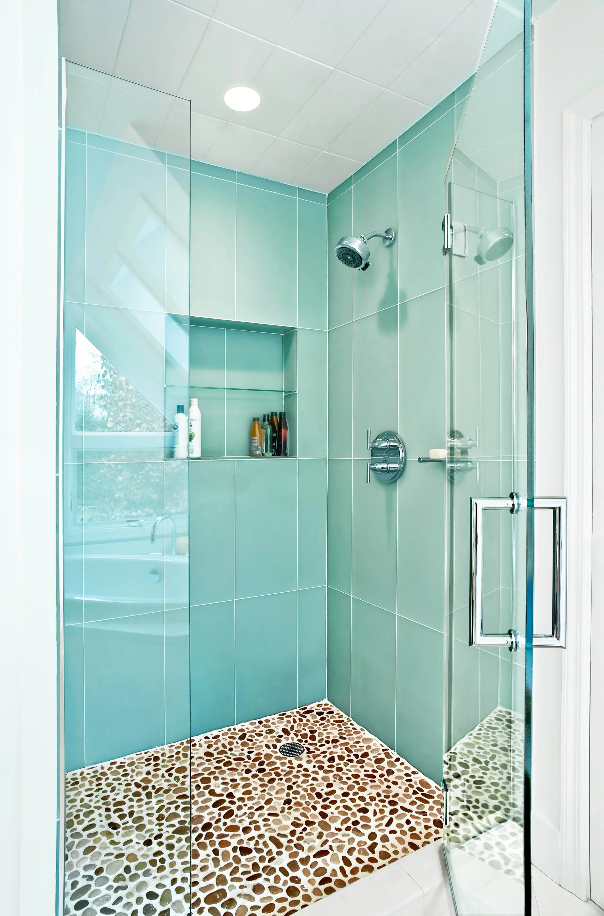 Walk-in shower with mint green glass tile and pebble stone floor in a Pleasantville, NY primary bathroom designed by Luminosus Designs.