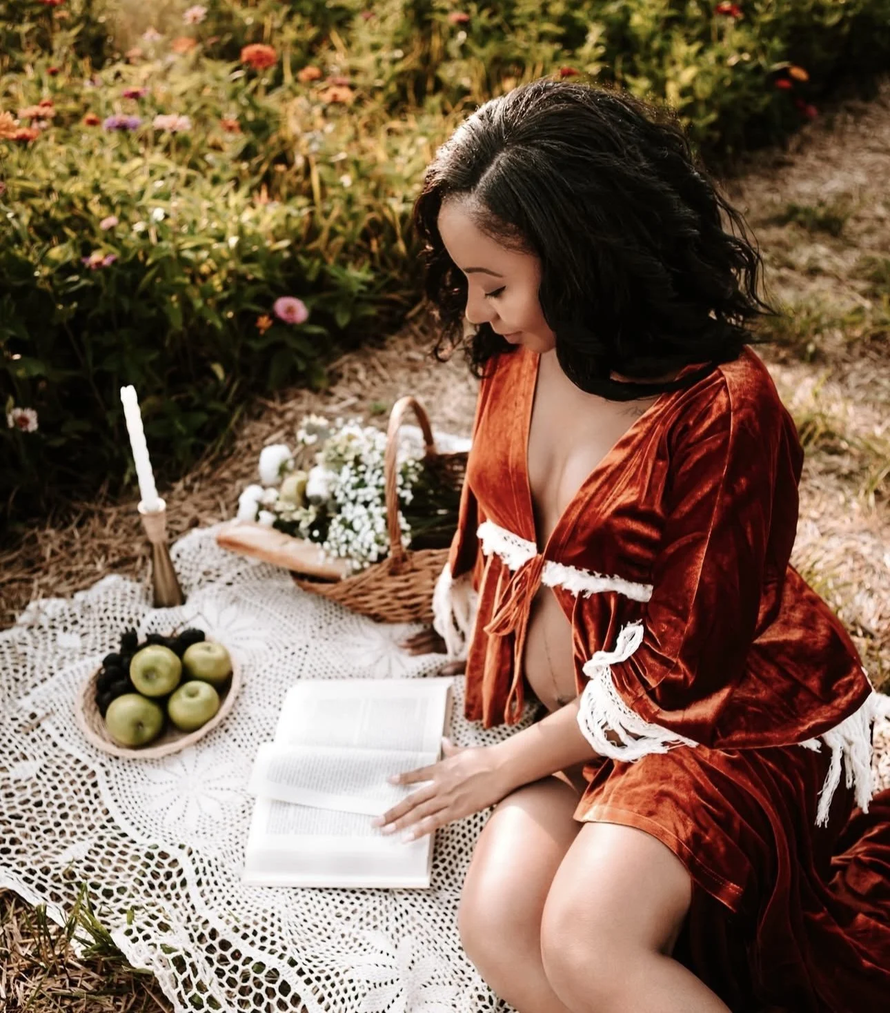 A woman in a velvet red dress sitting on a lace blanket outdoors, reading a book, surrounded by a picnic setup with apples, grapes, flowers, a candle, and a wicker basket, in a garden with colorful flowers.