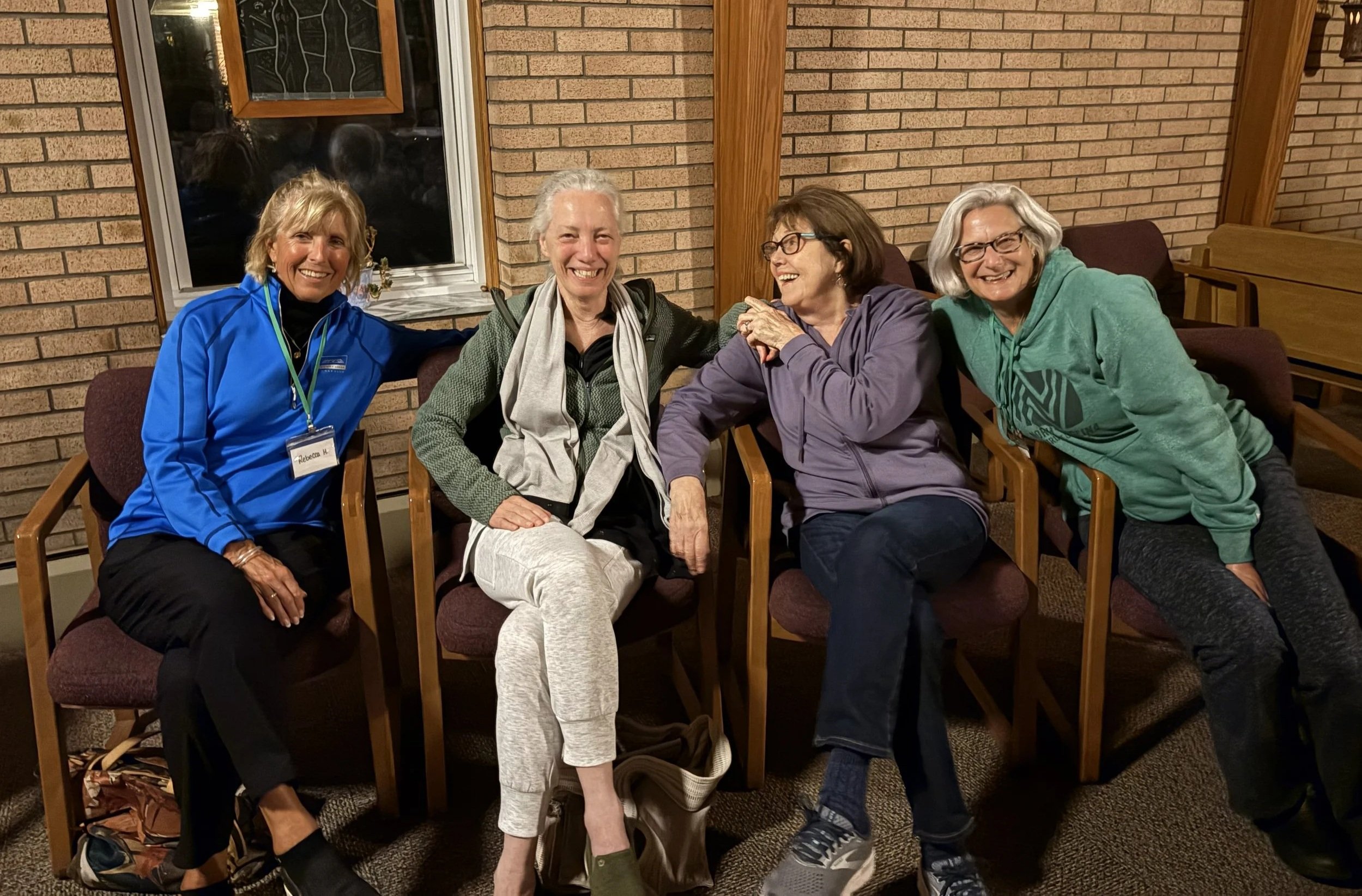 Four women sitting side-by-side on chairs, smiling and enjoying each other's company in an indoor setting with brick walls and wooden beams. Betty Hill Crowson The Joy is in the Journey Spiritual Retreats & Retreat Director Training