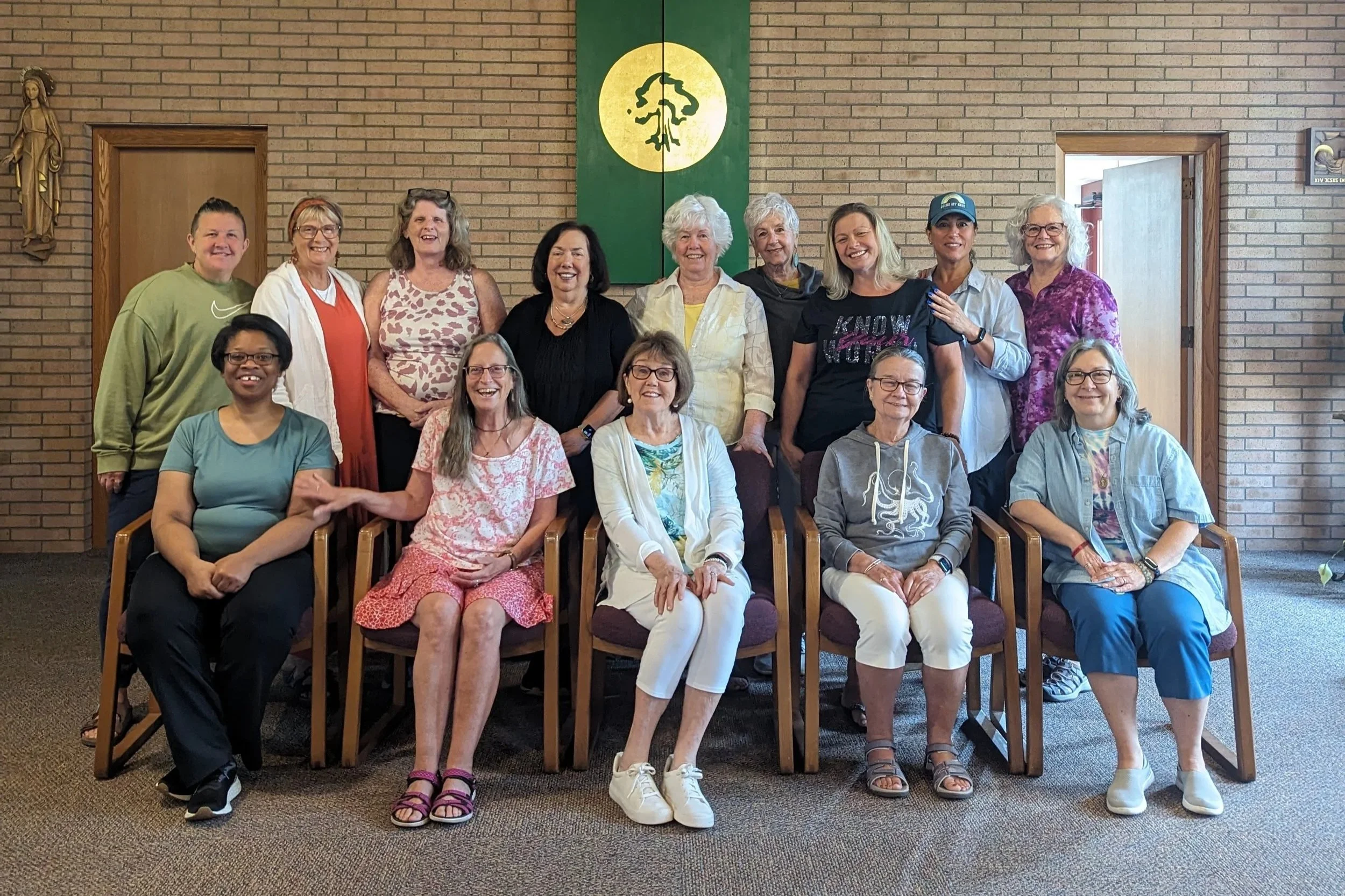 Group of fifteen women and one young man in a room with brick walls and Christian art, posing for a photo in front of a green and yellow religious banner. Betty Hill Crowson The Joy is in the Journey
