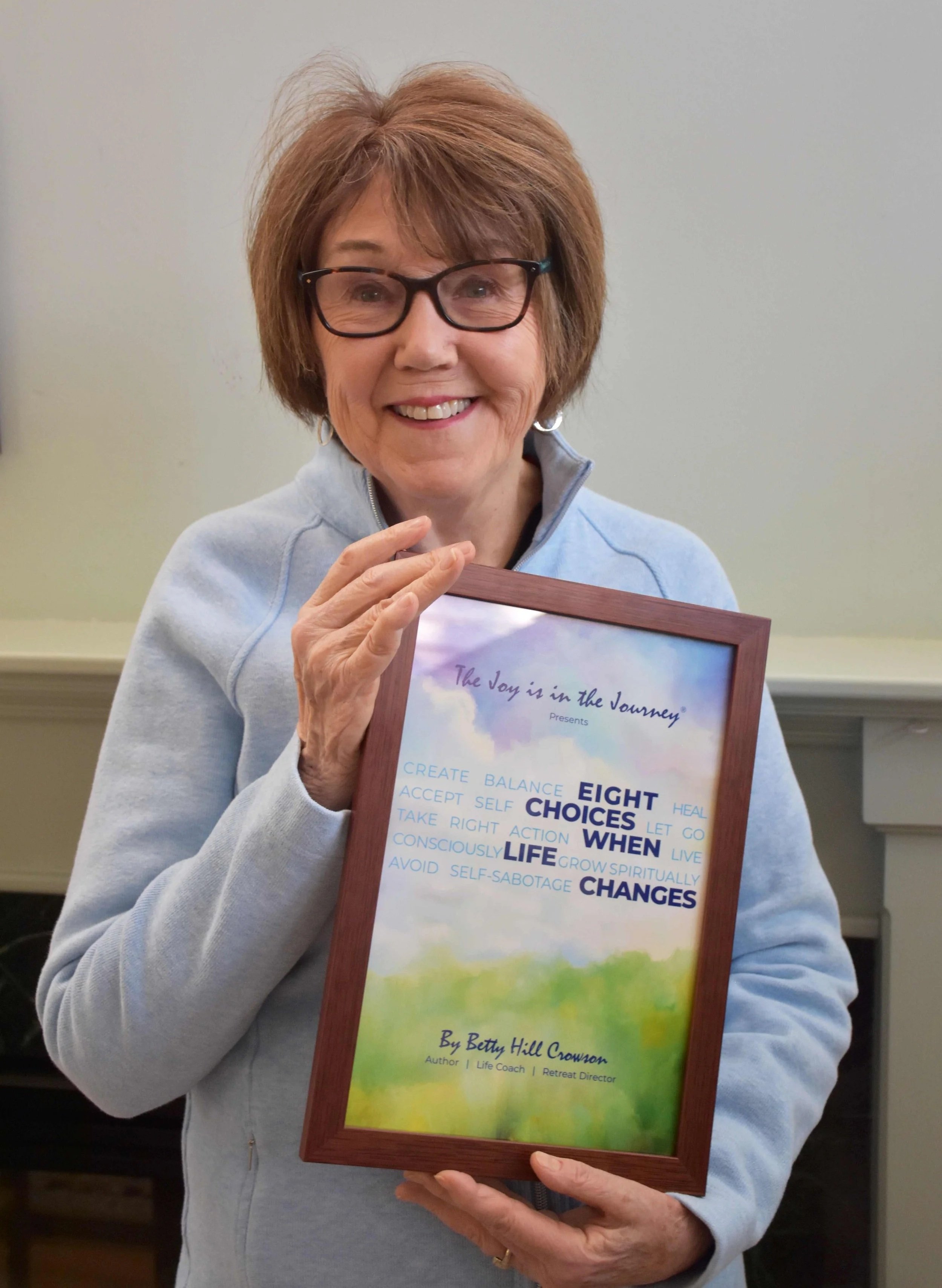 An older woman with glasses and short brown hair, smiling, holding a framed book titled 'The Joy is in the Journey' by Betty Hill Crowson, with a background of a neutral-colored wall. Betty Hill Crowson The Joy is in the Journey Spiritual Retreats &
