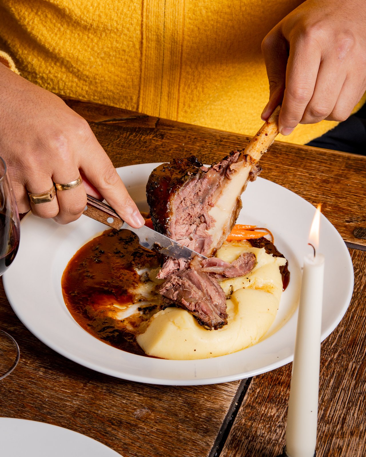 Person in a yellow sweater serves mashed potatoes with beef rib on a white plate, a candle on a wooden table.