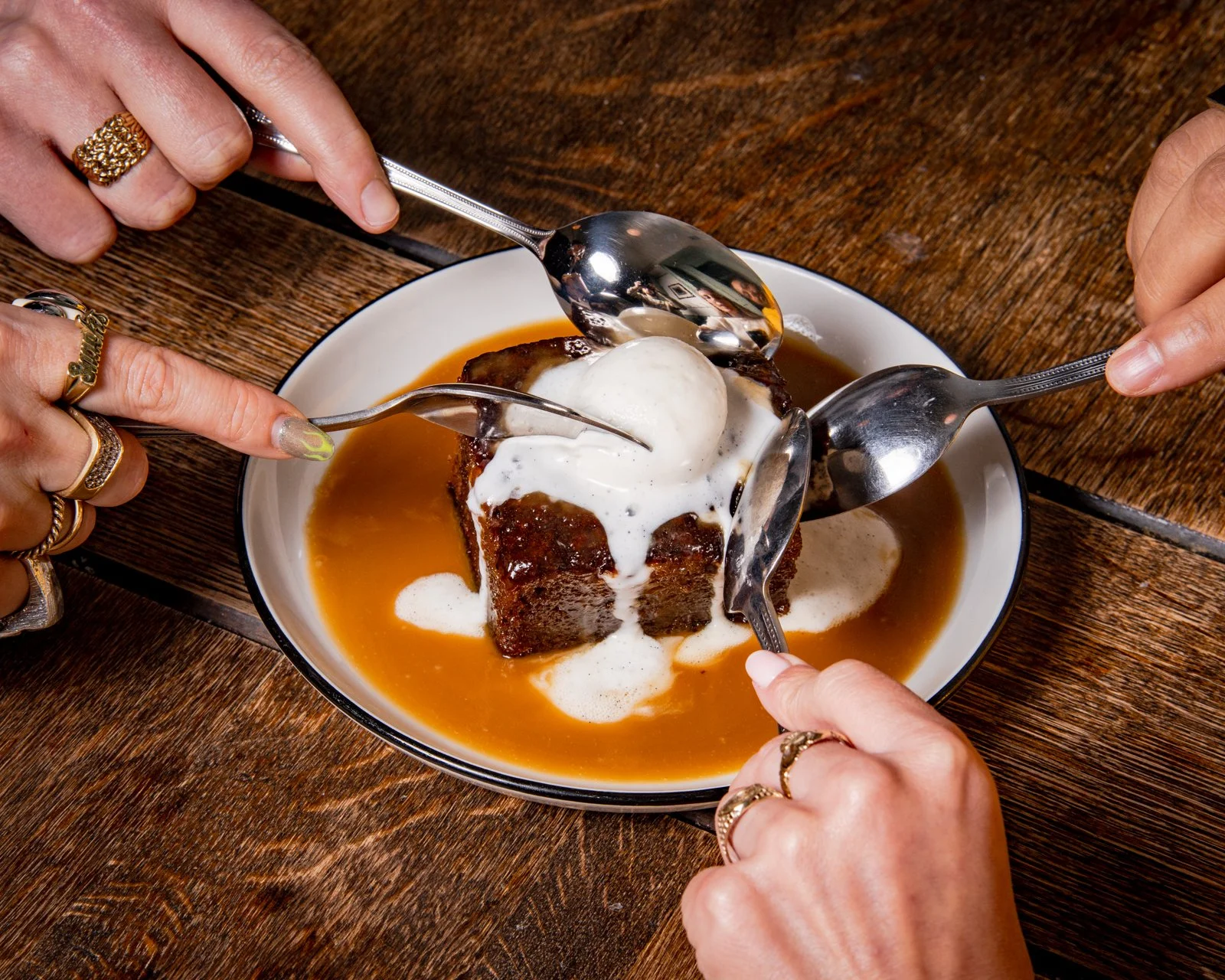 A dessert with a piece of chocolate cake topped with a scoop of vanilla ice cream, drizzled with caramel sauce, and topped with whipped cream. Four hands hold spoons around the plate to serve the dessert on a wooden table.