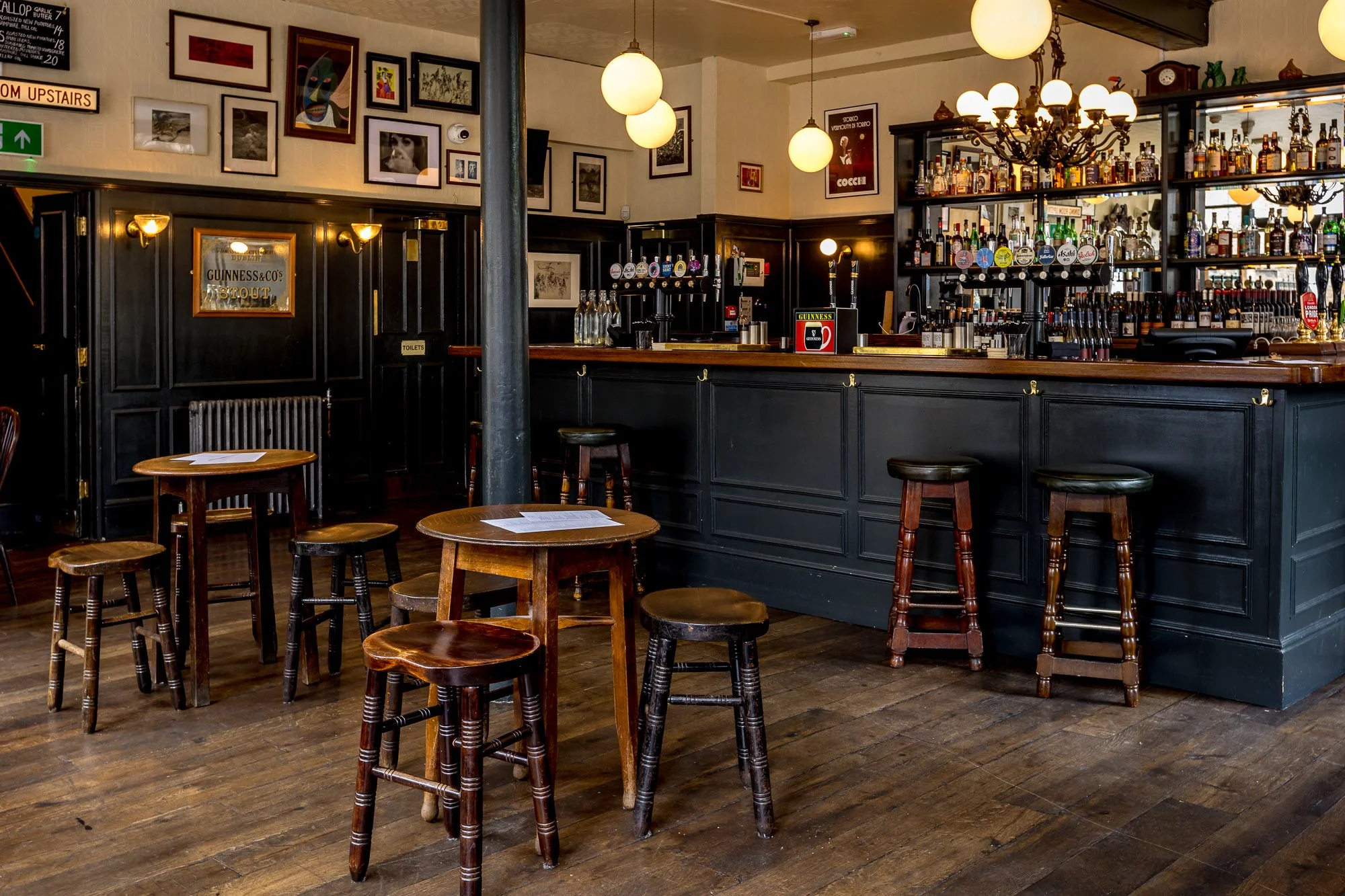 Interior of a pub or bar with a black wooden counter, various bottles of alcohol on shelves, hanging lights, wooden stools, and tables with chairs in a cozy, decorated setting.