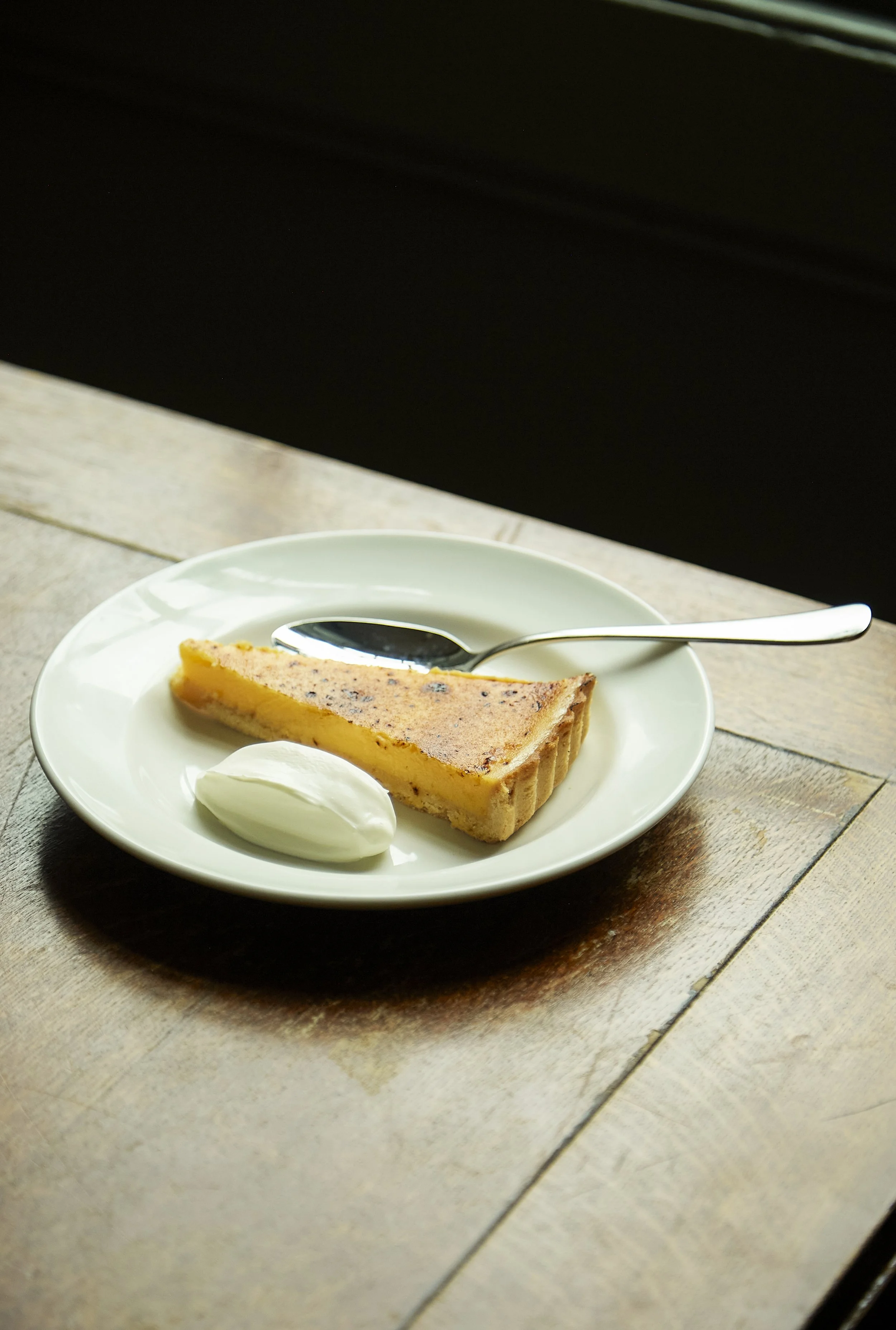 A slice of pumpkin pie with a dollop of whipped cream on a white plate, accompanied by a spoon, on a wooden table.