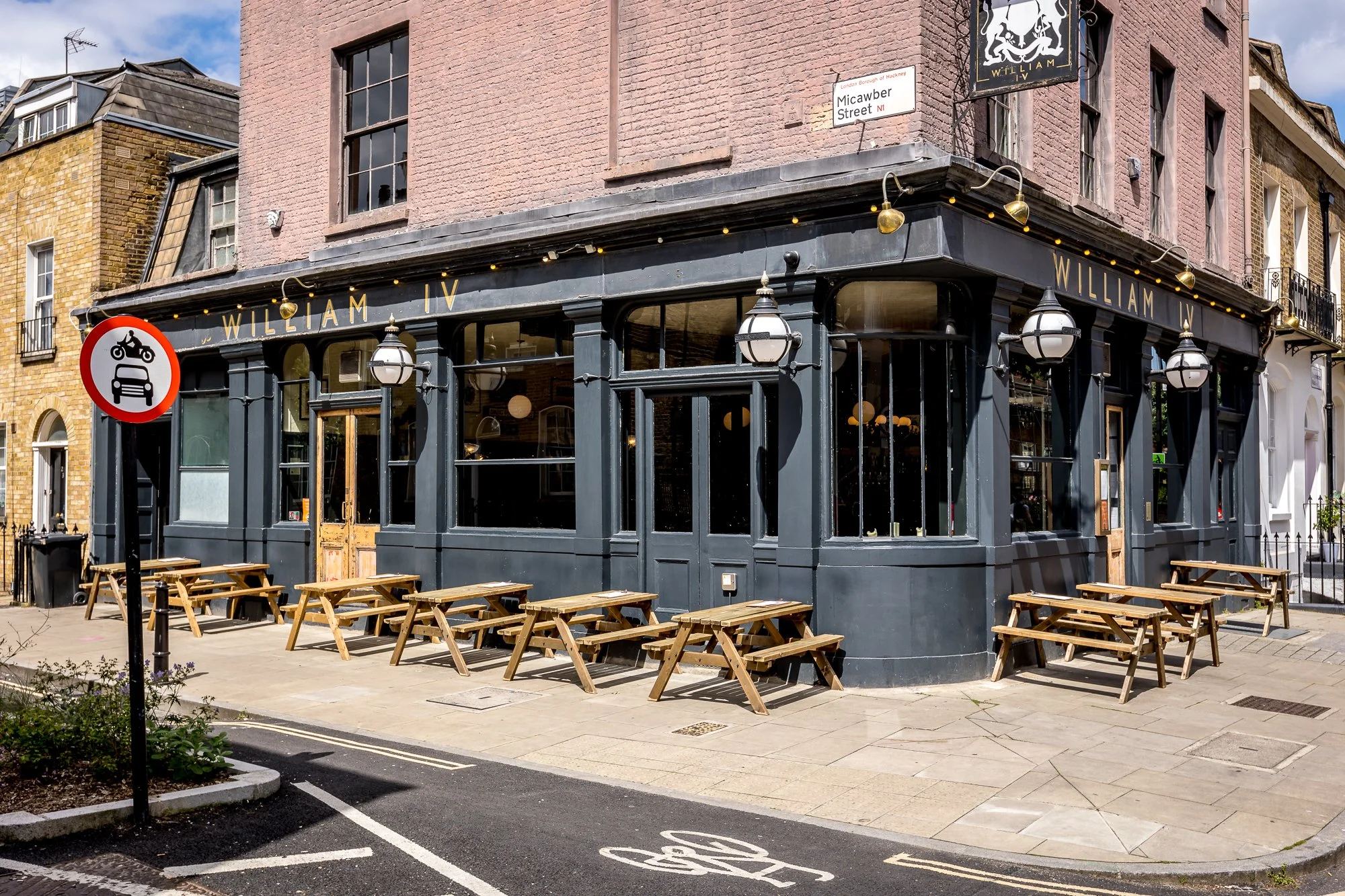 Outside view of a corner restaurant named William IV with wooden picnic tables on the sidewalk, a street sign for Micawber Street, and a no-entry sign for motorcycles and cars.