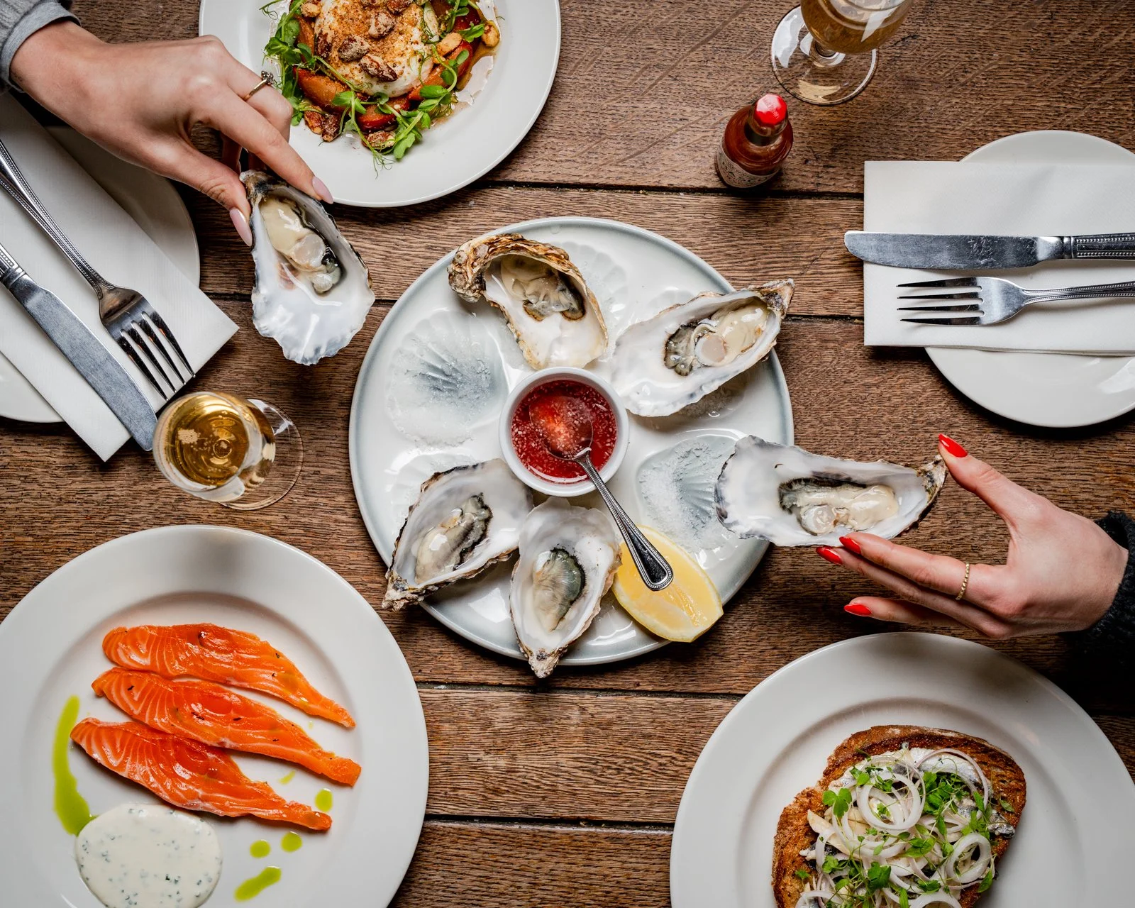 A top-down view of a dining table with plates of oysters, smoked salmon with sauce, and a salad with onions and microgreens. Two people are reaching for oysters, one with red painted nails, and there are glasses of wine, a bottle of hot sauce, and le