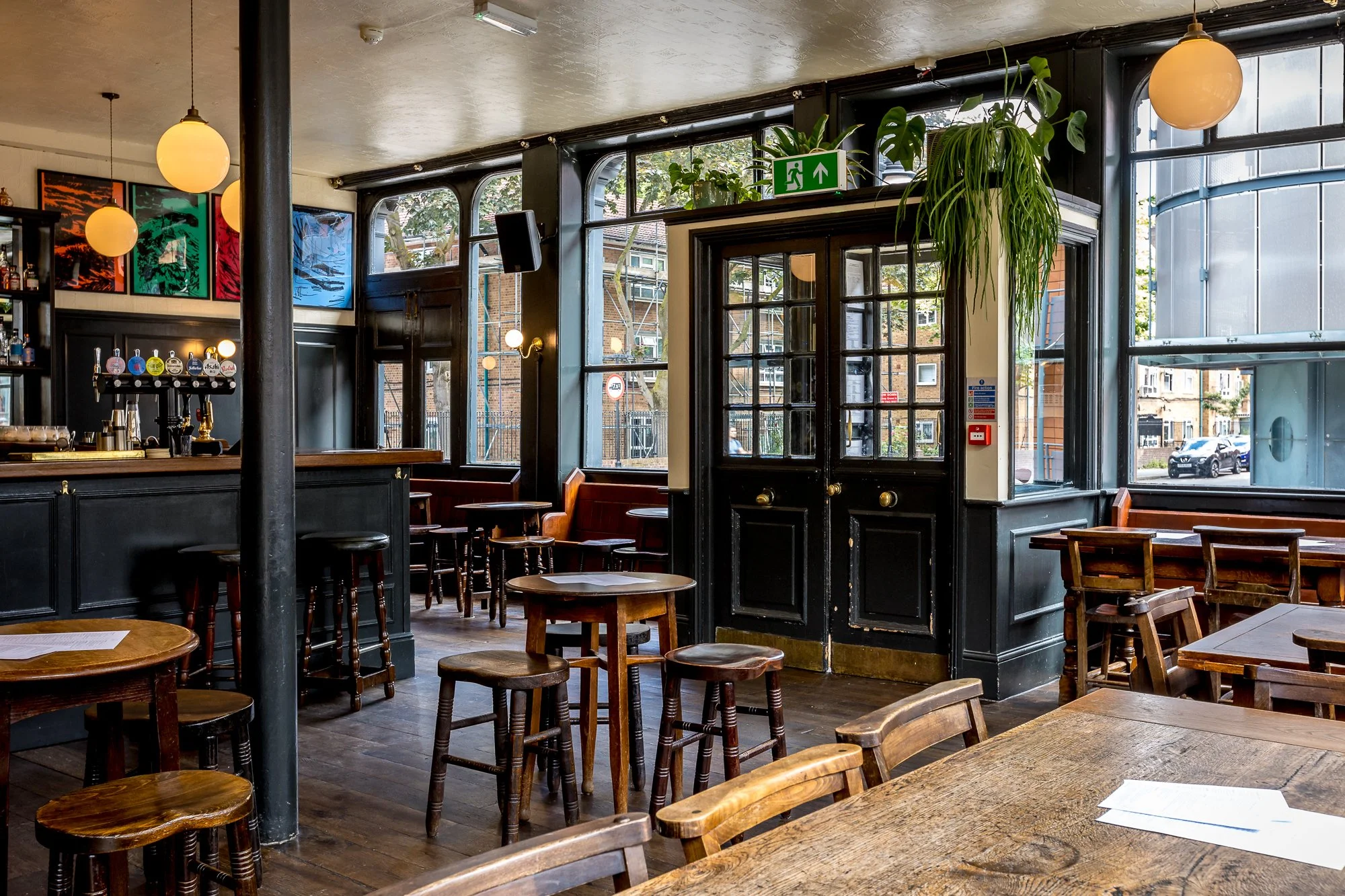 Interior of a cozy bar or pub with wooden tables and chairs, colorful artwork on the wall, large windows, and a bar area with taps.