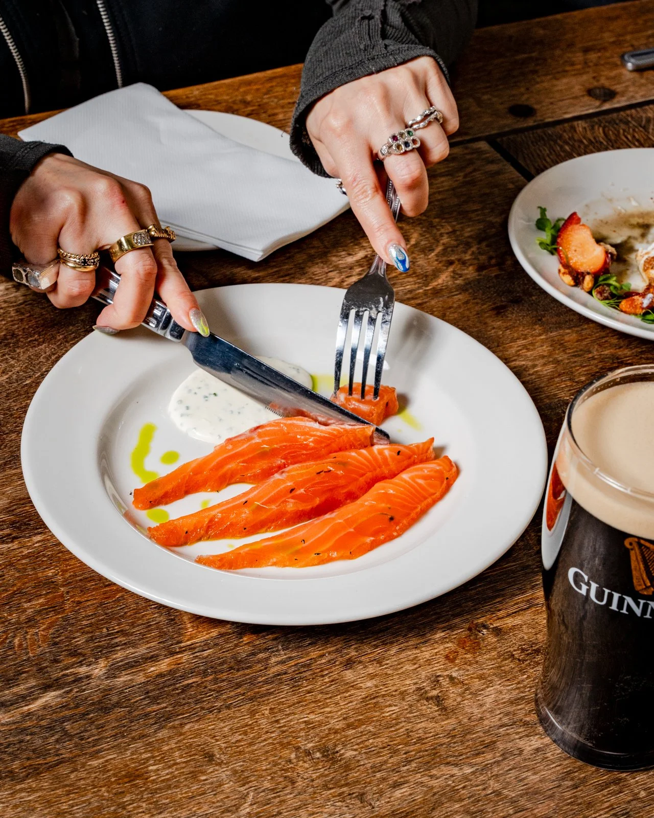 Person cutting smoked salmon with fork and knife at a wooden table with a plate of smoked salmon, a partially eaten salad plate, a Guinness beer, and a white napkin.