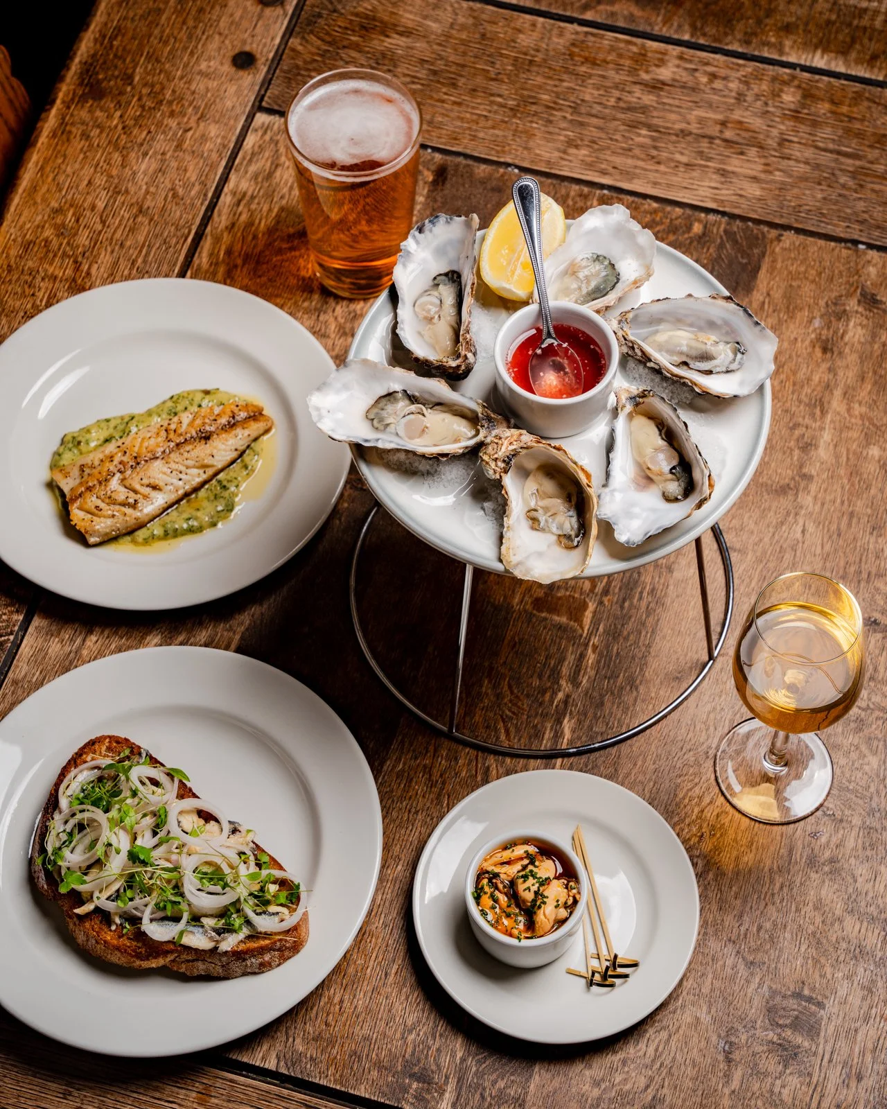 A wooden table with a plate of cooked salmon, a plate of toasted bread with onions and herbs, a glass of beer, a glass of white wine, and a seafood platter of oysters with lemon wedges and cocktail sauce.