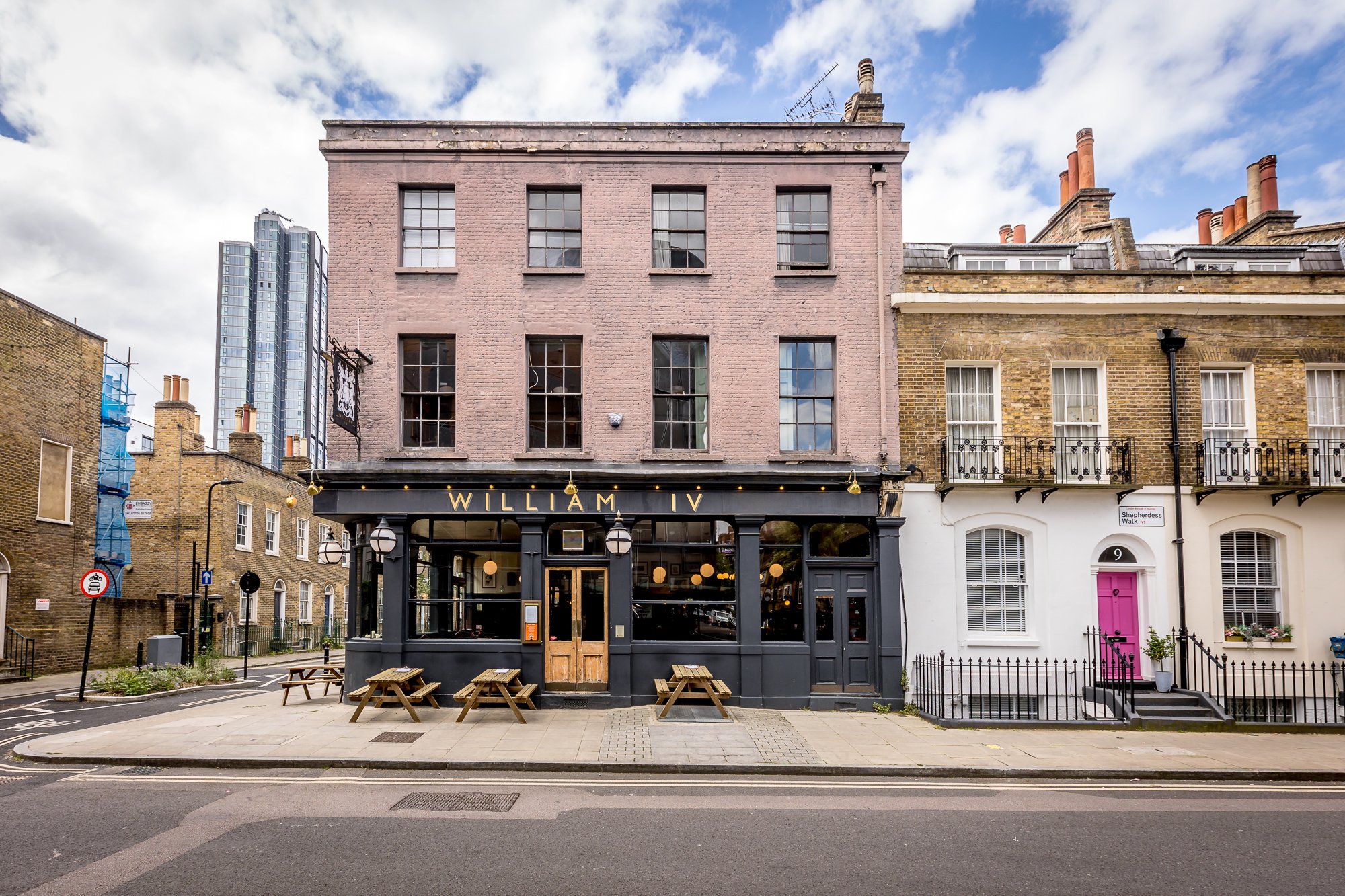 Front view of a building with a restaurant named William IV on the ground floor, with outdoor wooden benches, pink and yellow buildings, and a city street with a blue sky and clouds.