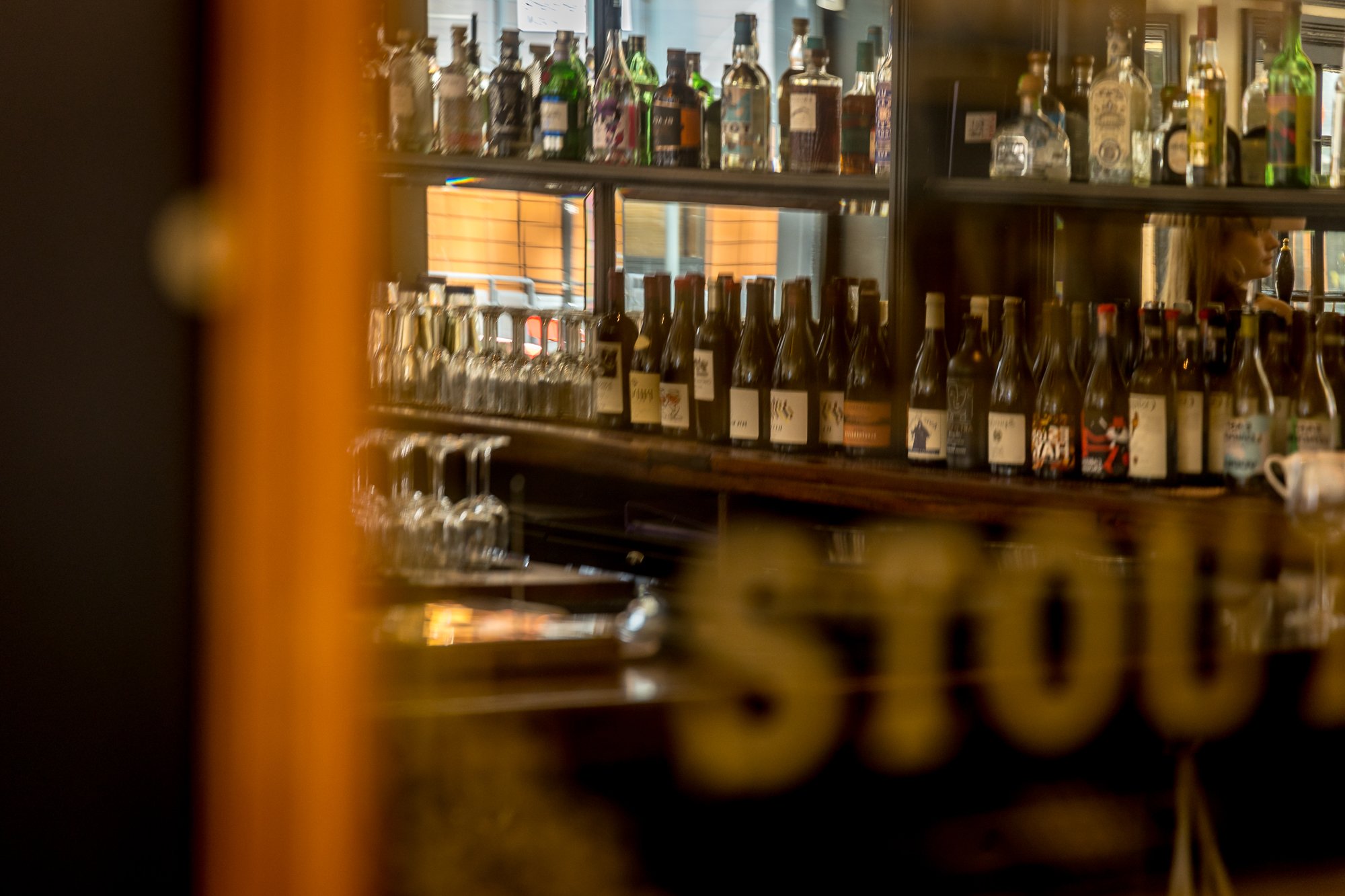 A bar with shelves of various bottles of alcohol, some with labels and glasses, viewed through a partially obscured window or mirror.