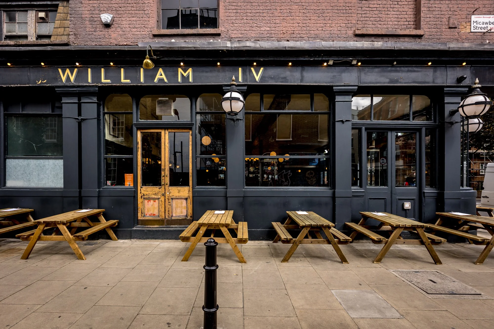 Exterior of a restaurant with outdoor wooden picnic tables, a black facade with gold lettering spelling 'WILLIAM IV,' and large glass windows, located on Micawber Street in London.