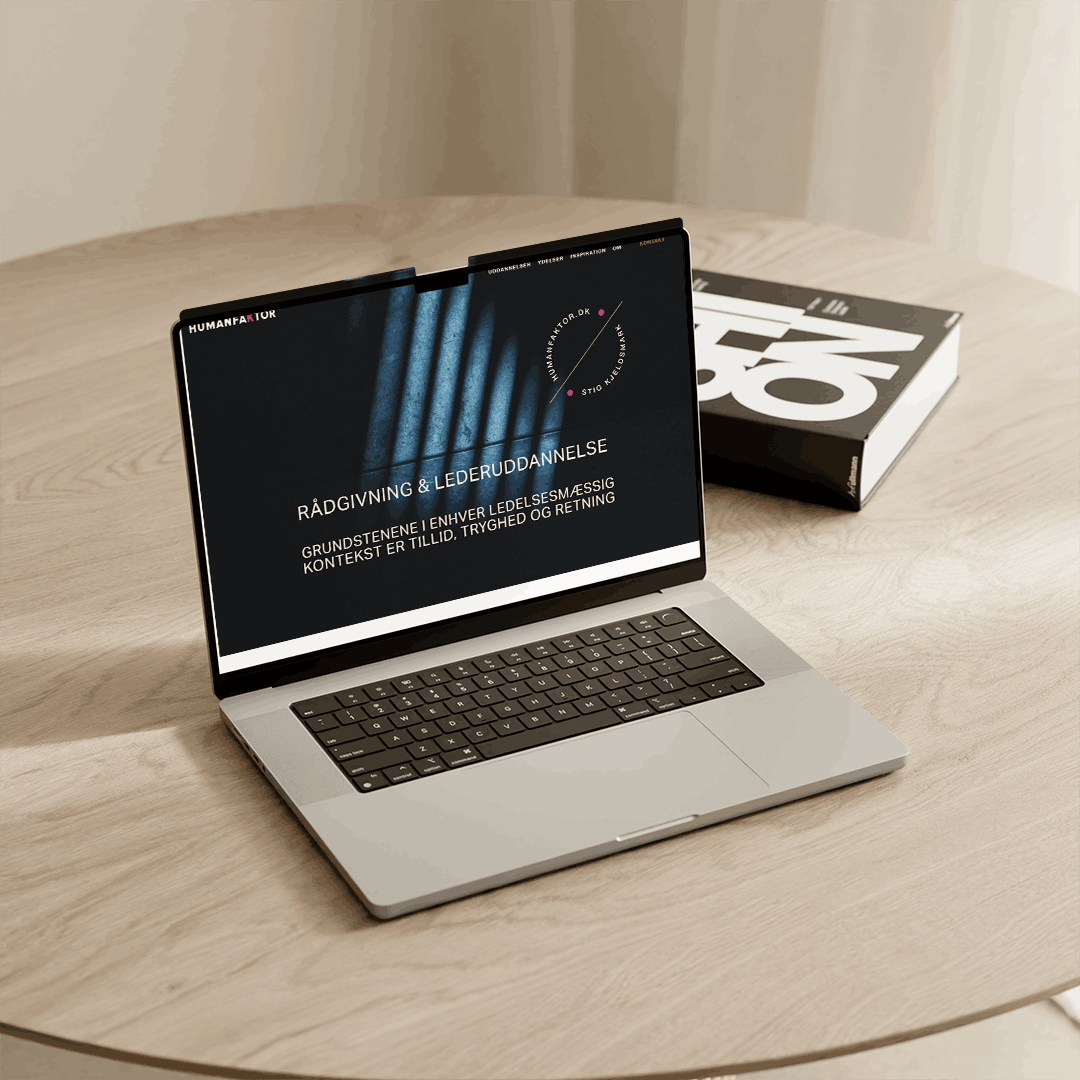 En bærbar computer,der  viser projekt designet af designcoach.dk  for humankaktor.dk, på et rundt træbord med en bog ved siden af. Skærmen viser hjemmesidens forside med titlen 'Rådgivning & Lederuddannelse'