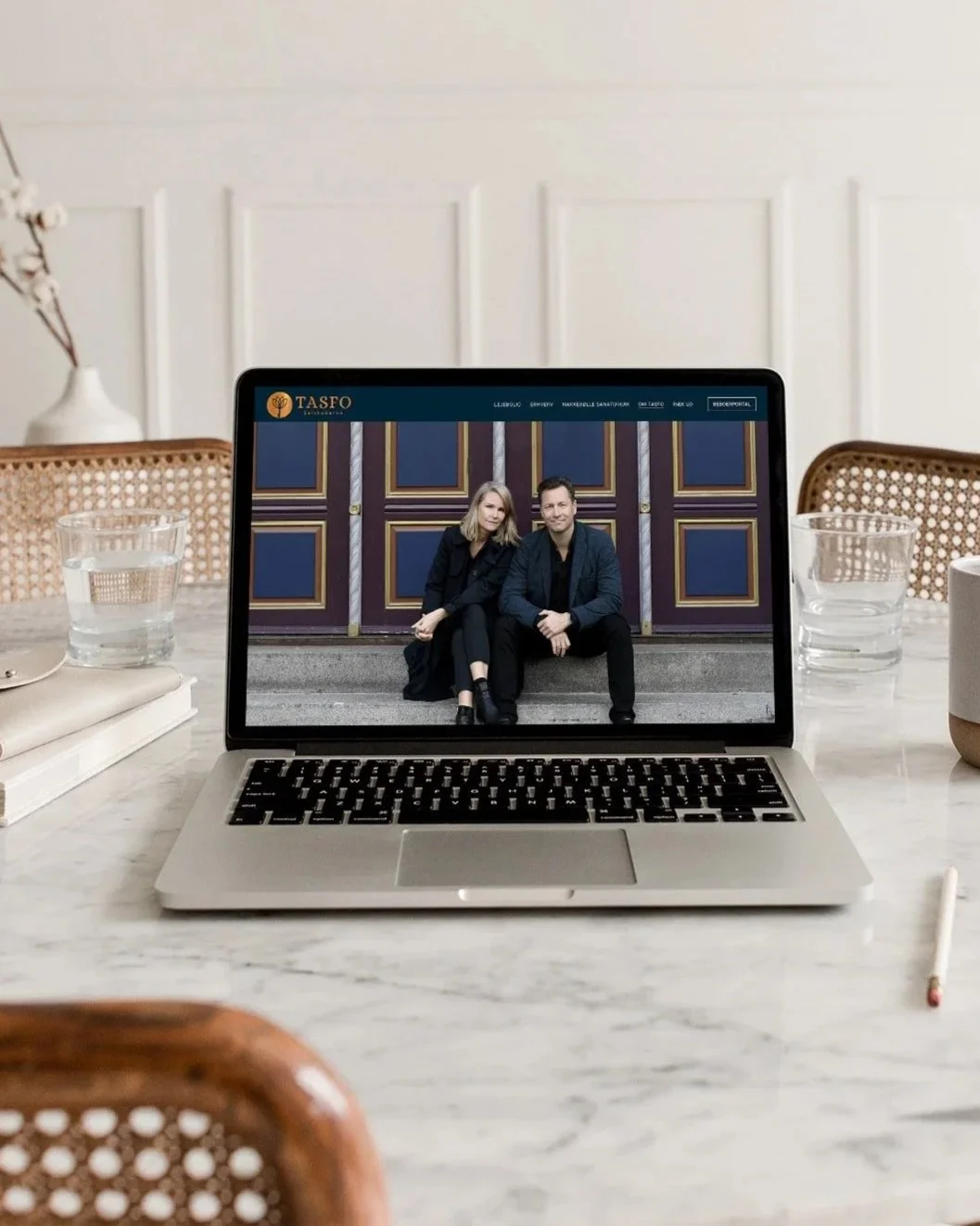En åben bærbar computer på et marmorbord viser en hjemmeside med et par siddende foran en dekorativ blå dør, på en indendørs setting.