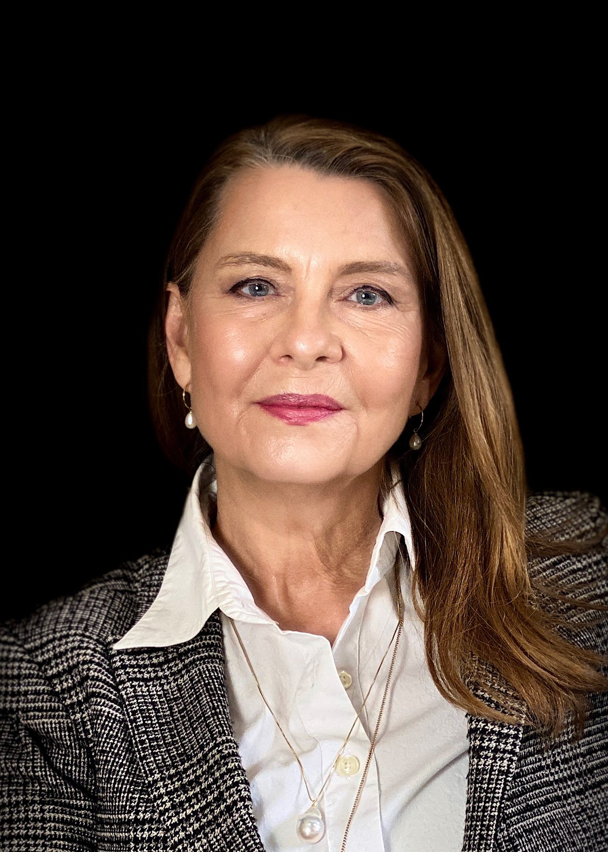 Portræt af Angela Lund. designcoach med brunt langt hår, blå øjne, iført hvid skjorte, tweed jakke og perleøreringe mod sort baggrund.