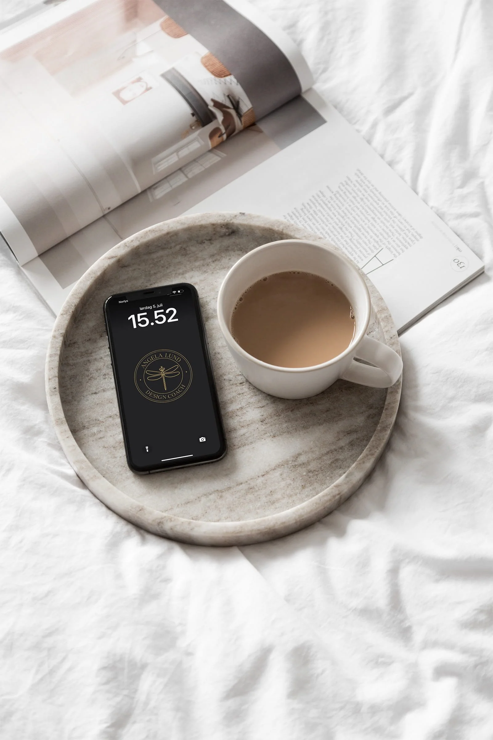 En rund marmorbakke med en kop kaffe og en smartphone liggende på en seng med et åbent magasin. Der er også en åben bog i baggrunden.