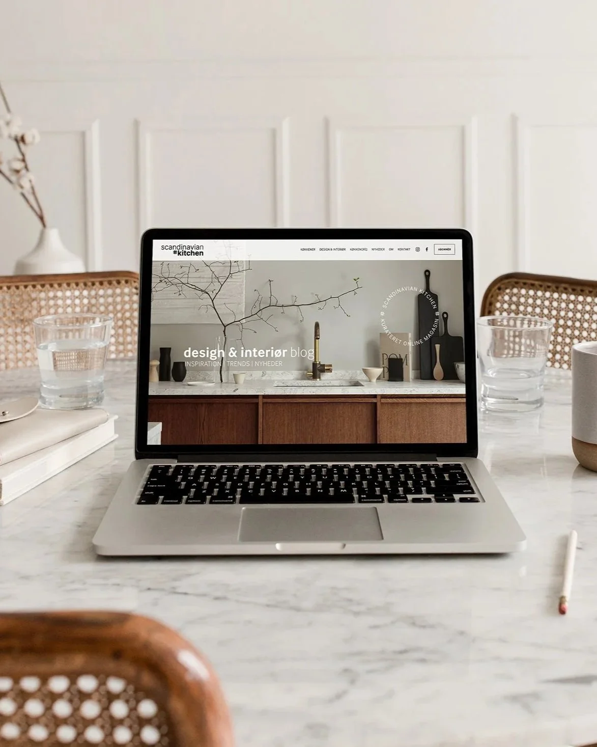 En bærbar computer, der viser en blog designet af Angela Lund -designcoach.dk - på et marmorbord med to glas vand og en notesbog. Baggrunden viser en hvid væg med træpaneler.